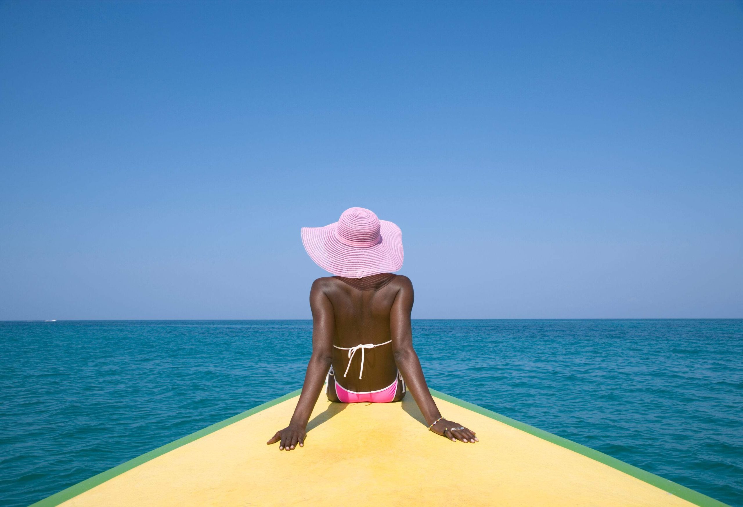 A dark-skinned female tourist in a pink bikini and hat sits on the boat's bow on the deep blue sea.