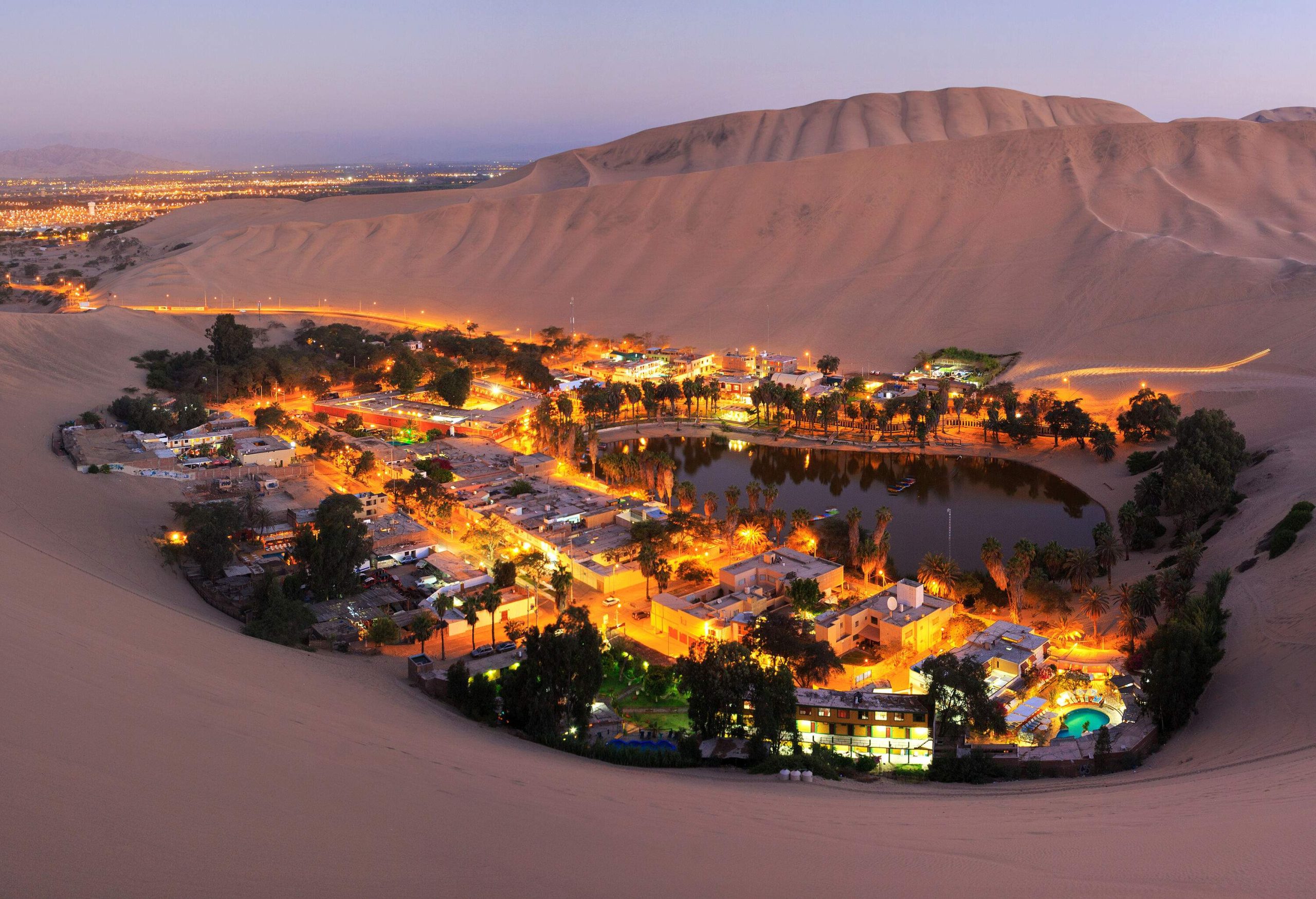A night-time view of a bright village in a desert oasis bordered by sand dunes.