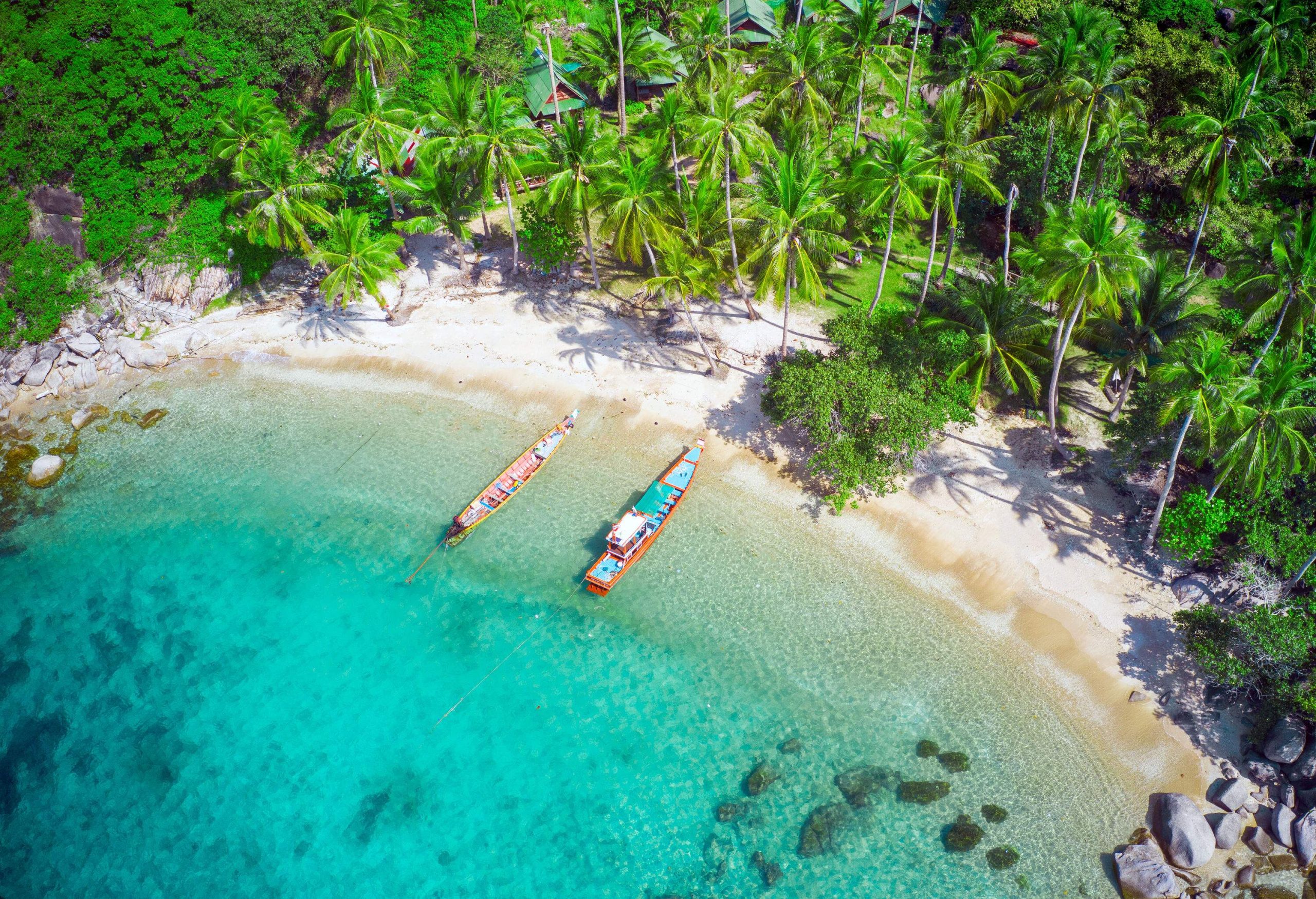 Aerial view of a turquoise crystal clear water beach with two long boats moored by the white sand shore surrounded by tall green trees.