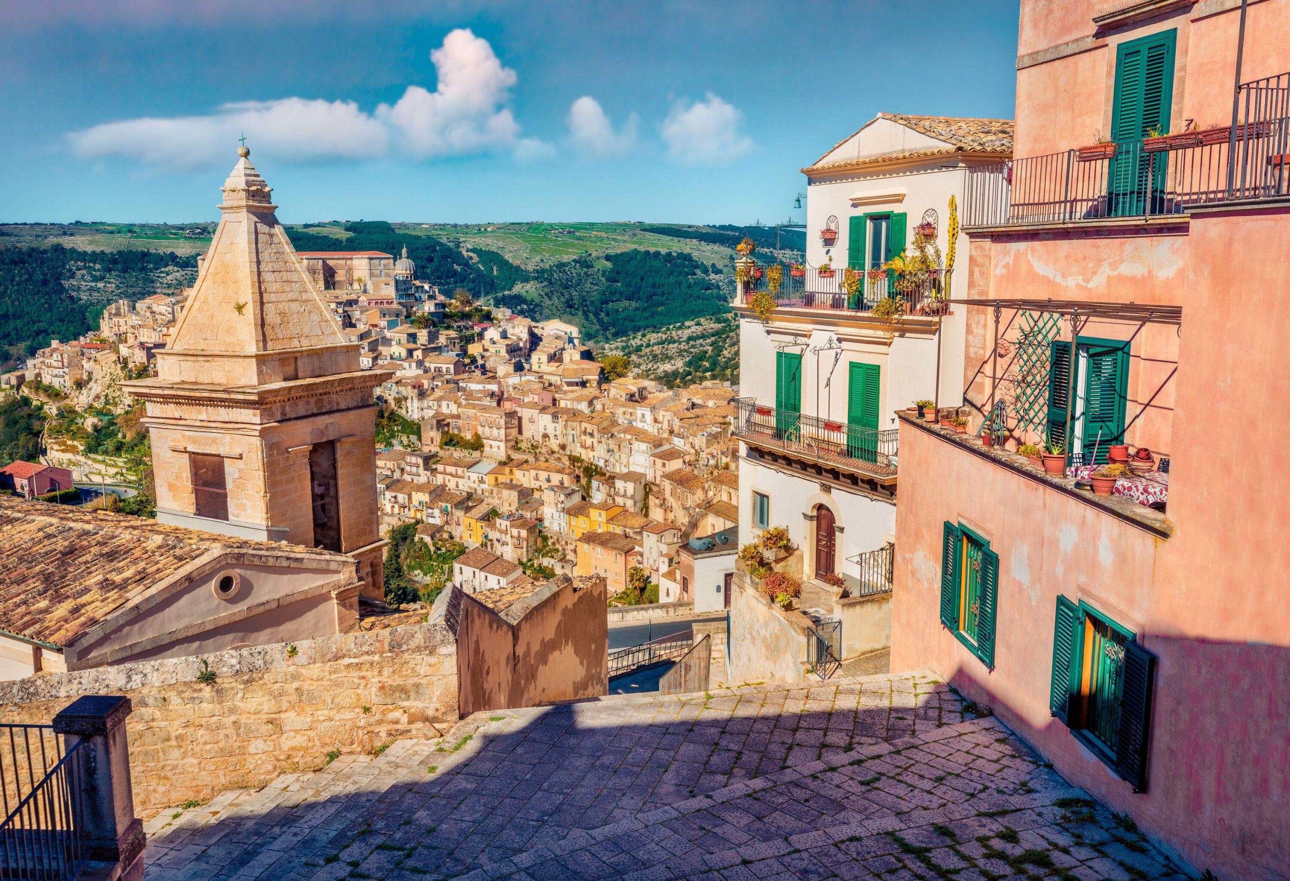 A hilltop staircase along a church steeple and terraced buildings overlooking a cluster of buildings perched on hills.