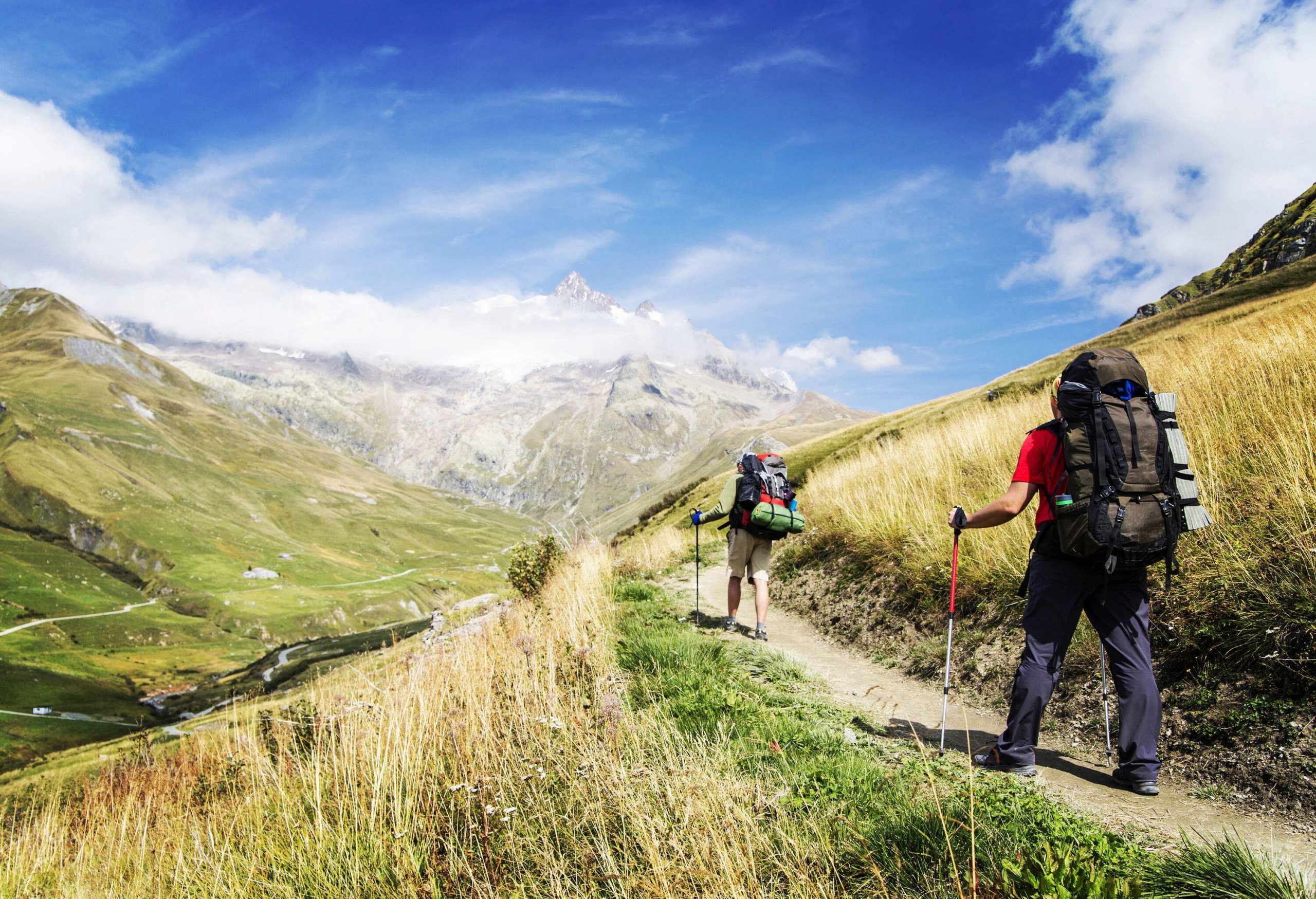 Two hikers carrying camping gear travel down a grassy hillside with distant vistas of lush valleys and cloud-covered mountains.