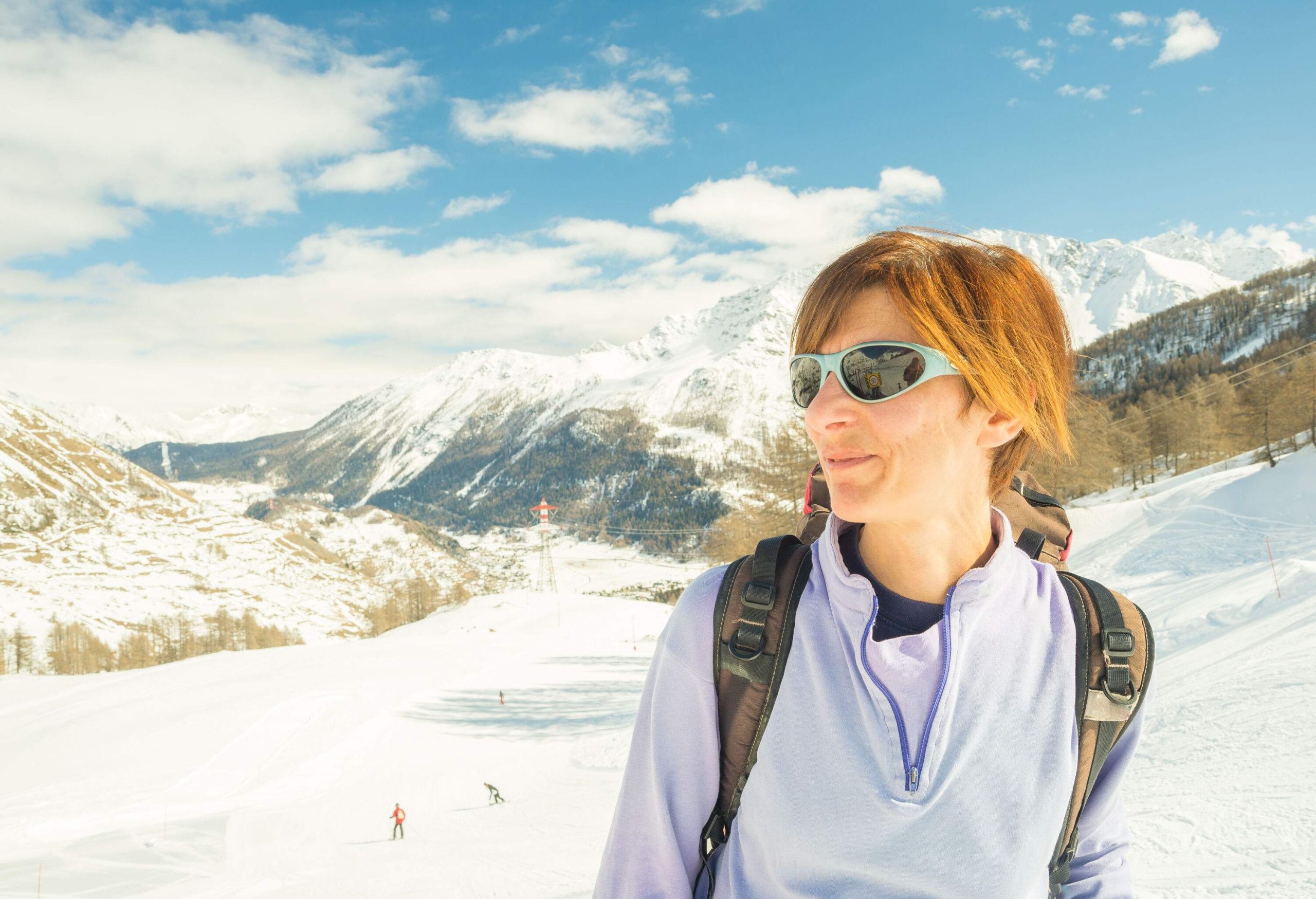A young woman with a backpack and sunglasses standing in the middle of a ski slope.