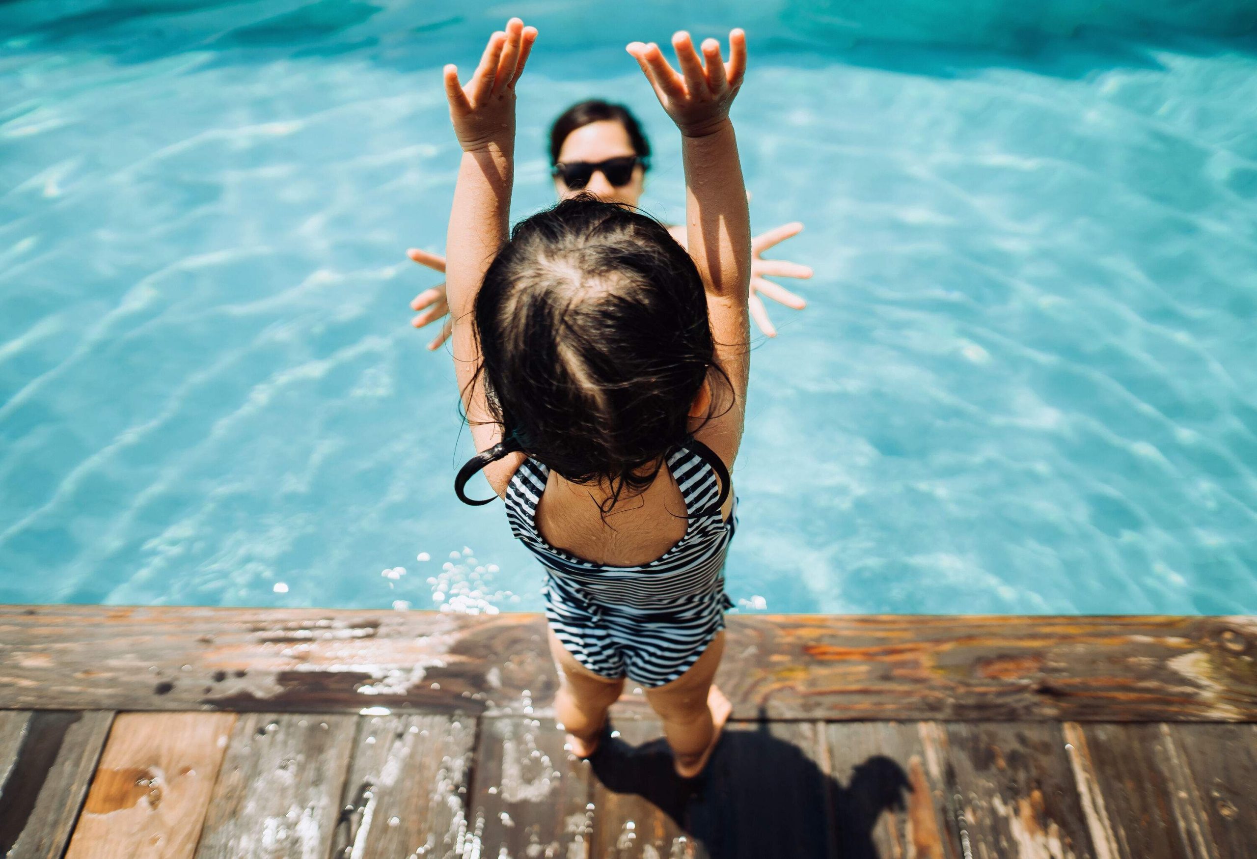 An excited toddler girl stands poolside with her arms raised as she watches her mother in the water.