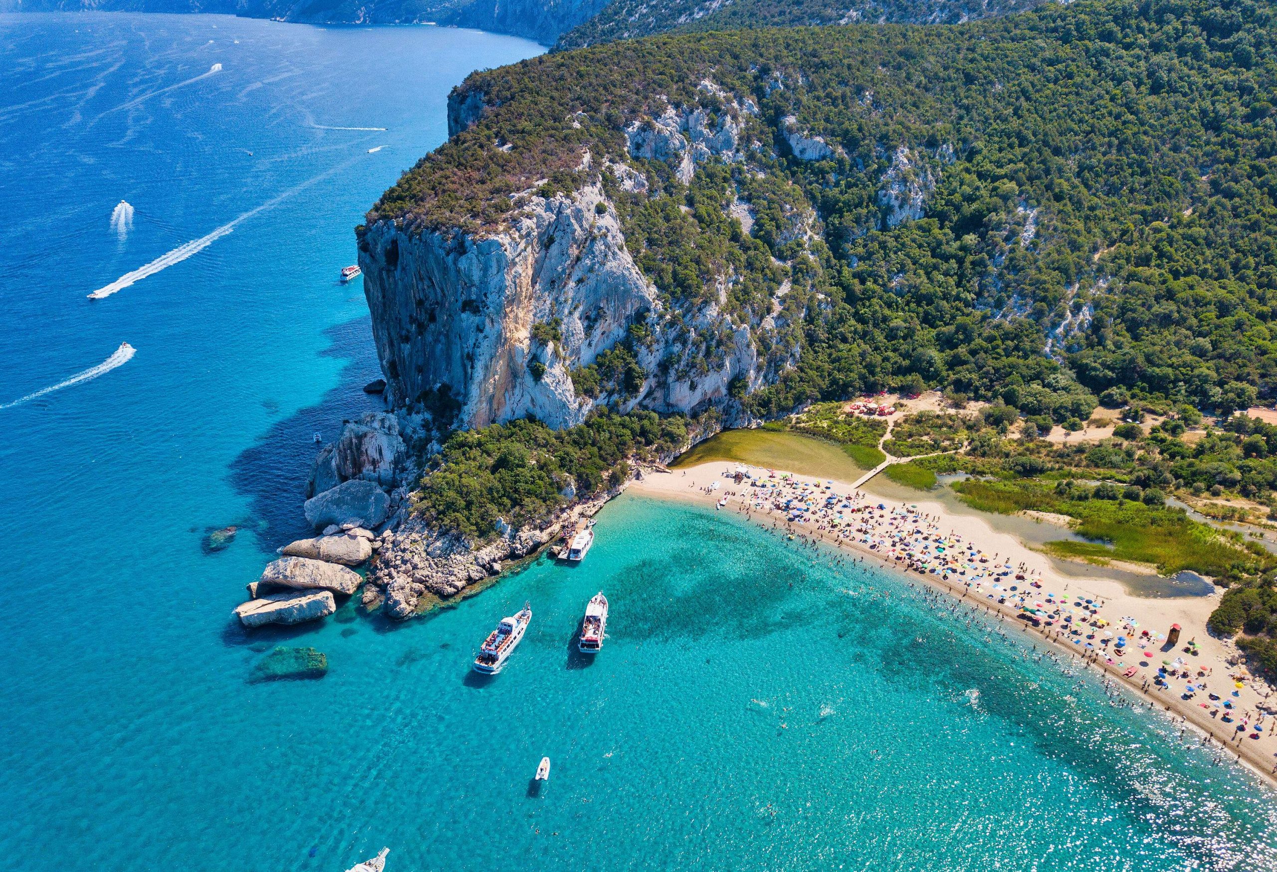 Aerial view of a lush cliff and a shore lined with colourful sunbeds and parasols by the turquoise sea.