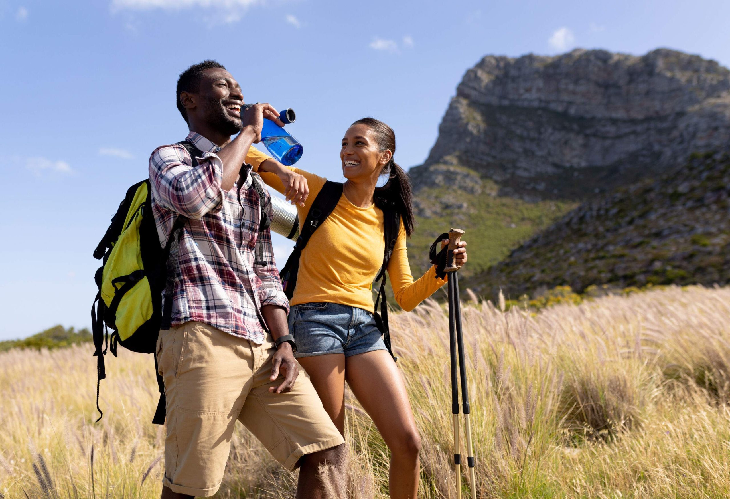 Two people strolling through tall grass with a big rock mountain behind them.