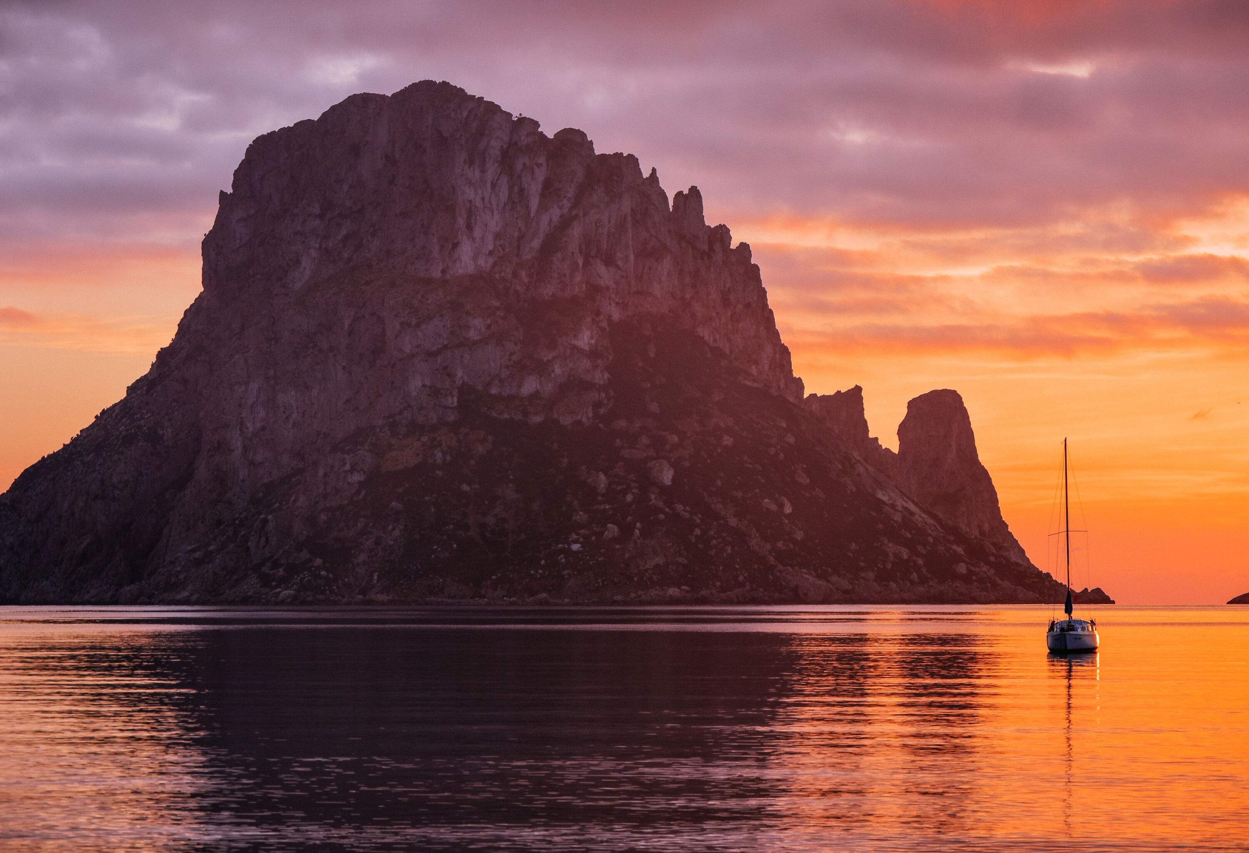 A small boat sailing on calm waters near a steep rocky mountain in the middle of a sea at sunset.