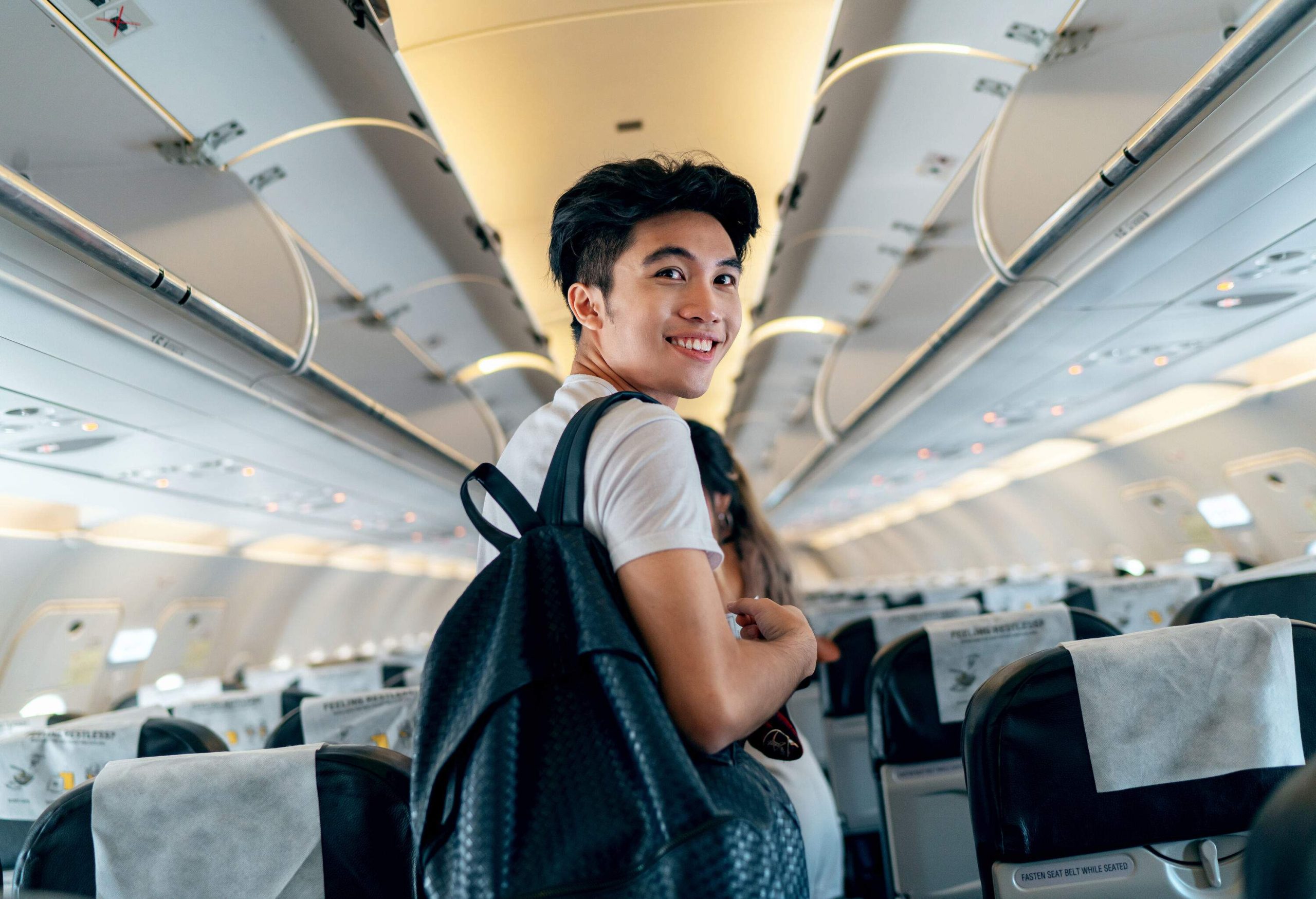 A young Asian male traveller boards a plane, carrying a backpack and glancing back.
