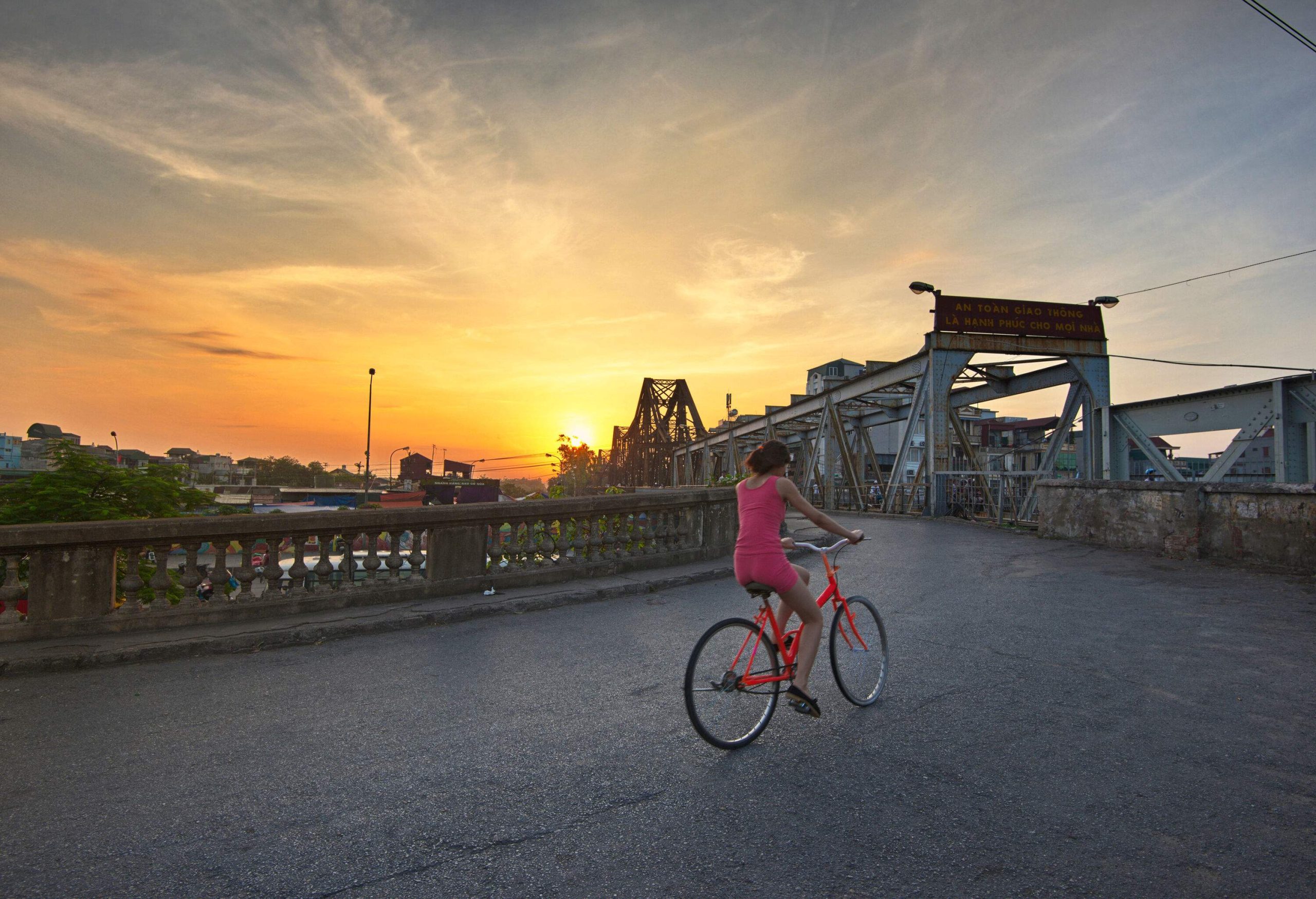 A woman rides a bicycle across a path beside a bridge with views of a beautiful sunset.