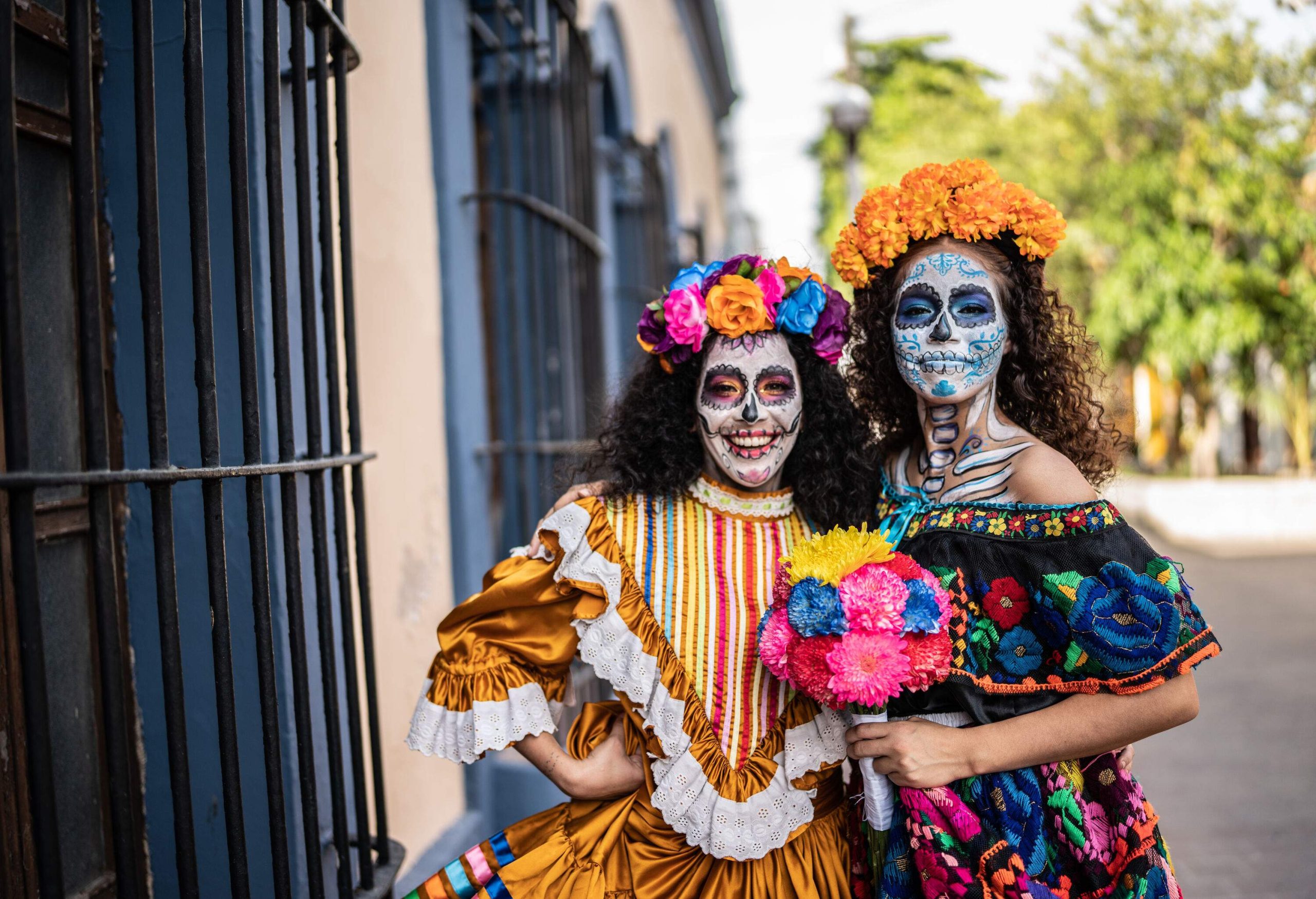 Two individuals in colourful dresses and flower headbands with their faces painted as skulls.