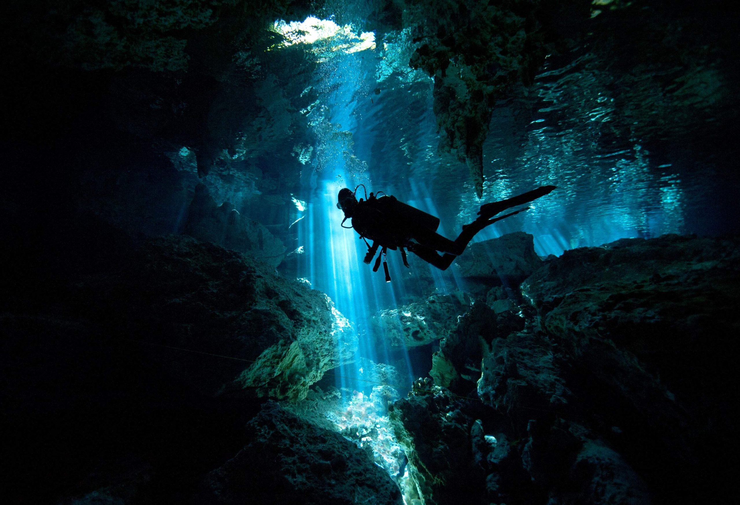 Silhouette of a scuba diver exploring an underwater cave with light streaks from small openings.