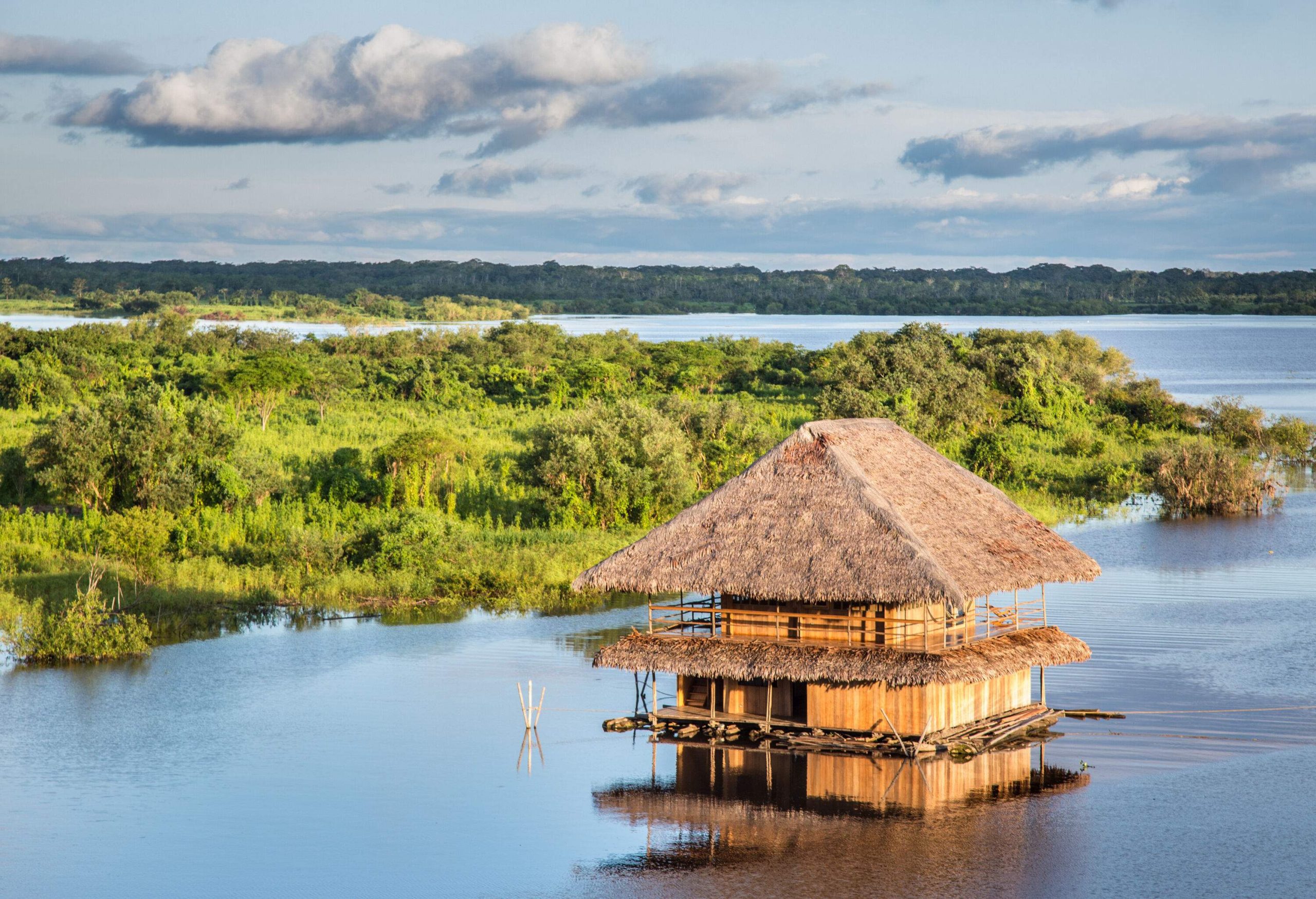 A wooden house with thatched roofs perched in the waters of a calm river with views of a grassy riverbank.