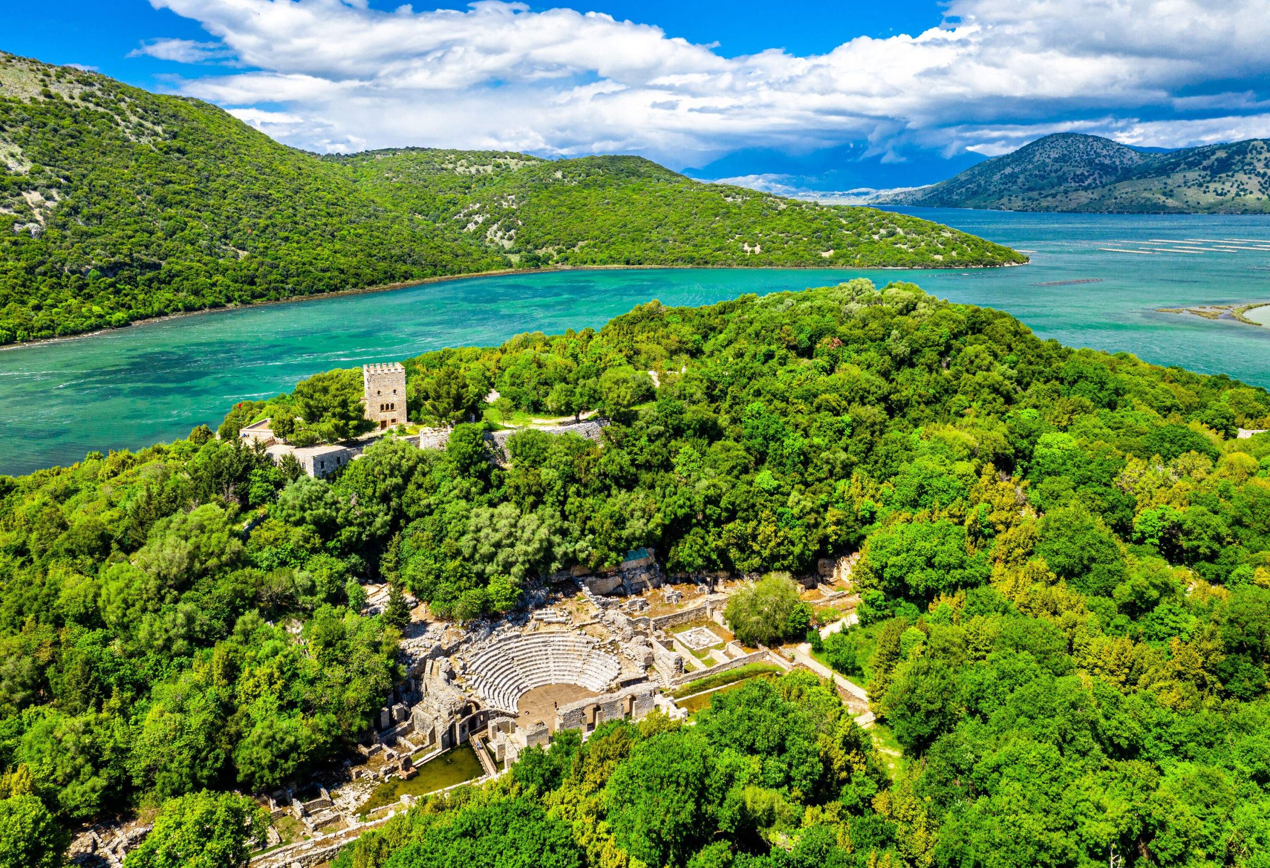 An archaeological site with ruins of ancient buildings and amphitheatre surrounded by a dense forest beside a lake.