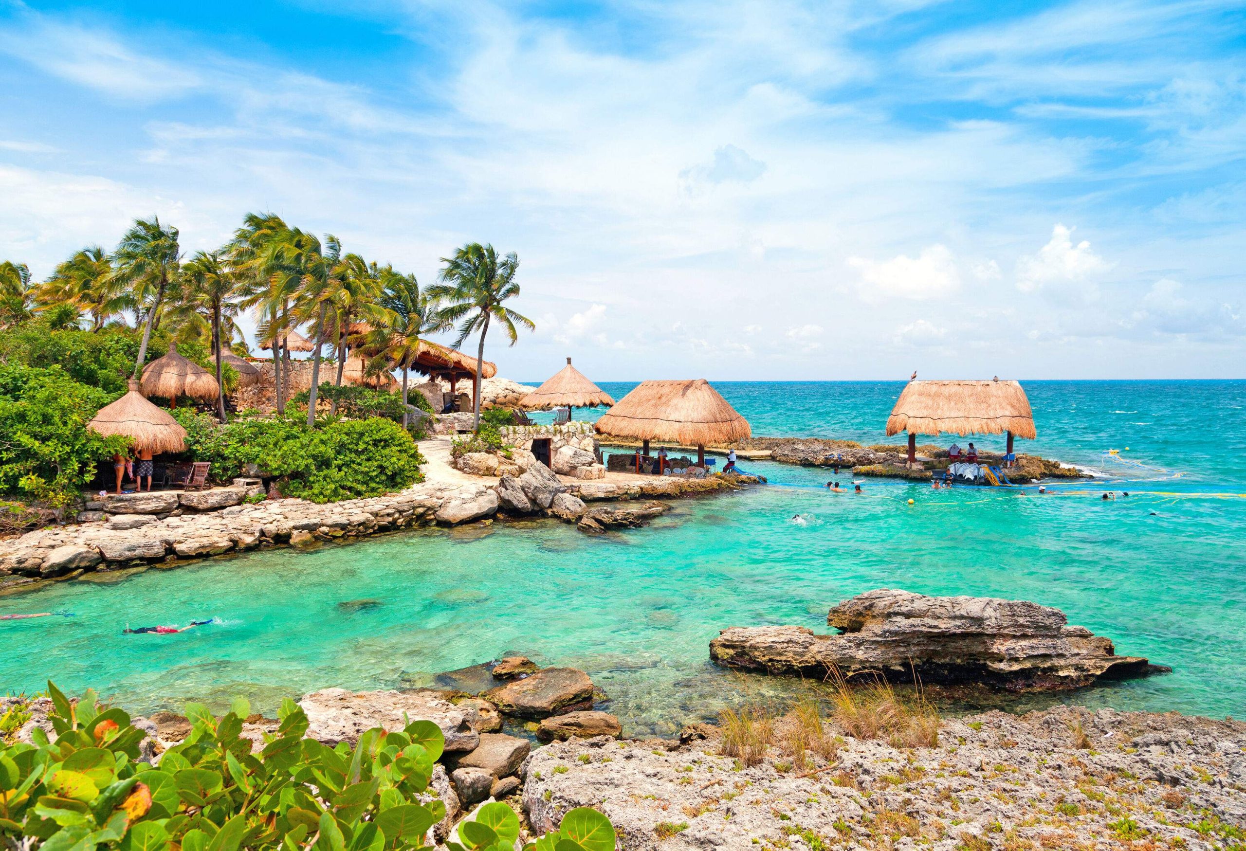 Wooden beach huts on the rocky shore surrounded by tall green trees by the blue beach.