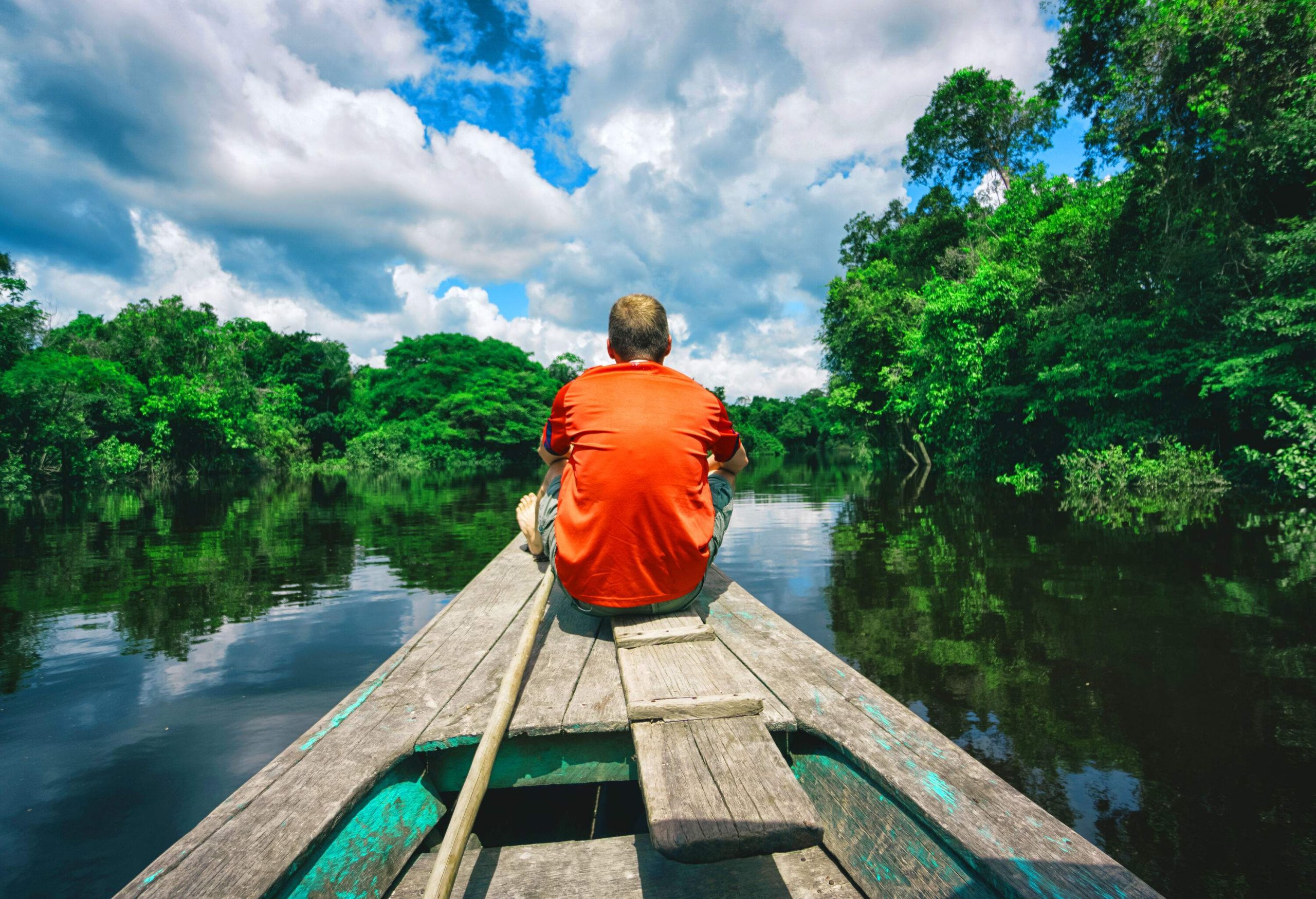 A man wearing an orange t-shirt sits at the bow of a wooden boat on a river surrounded by verdant trees against the cloudy blue sky.