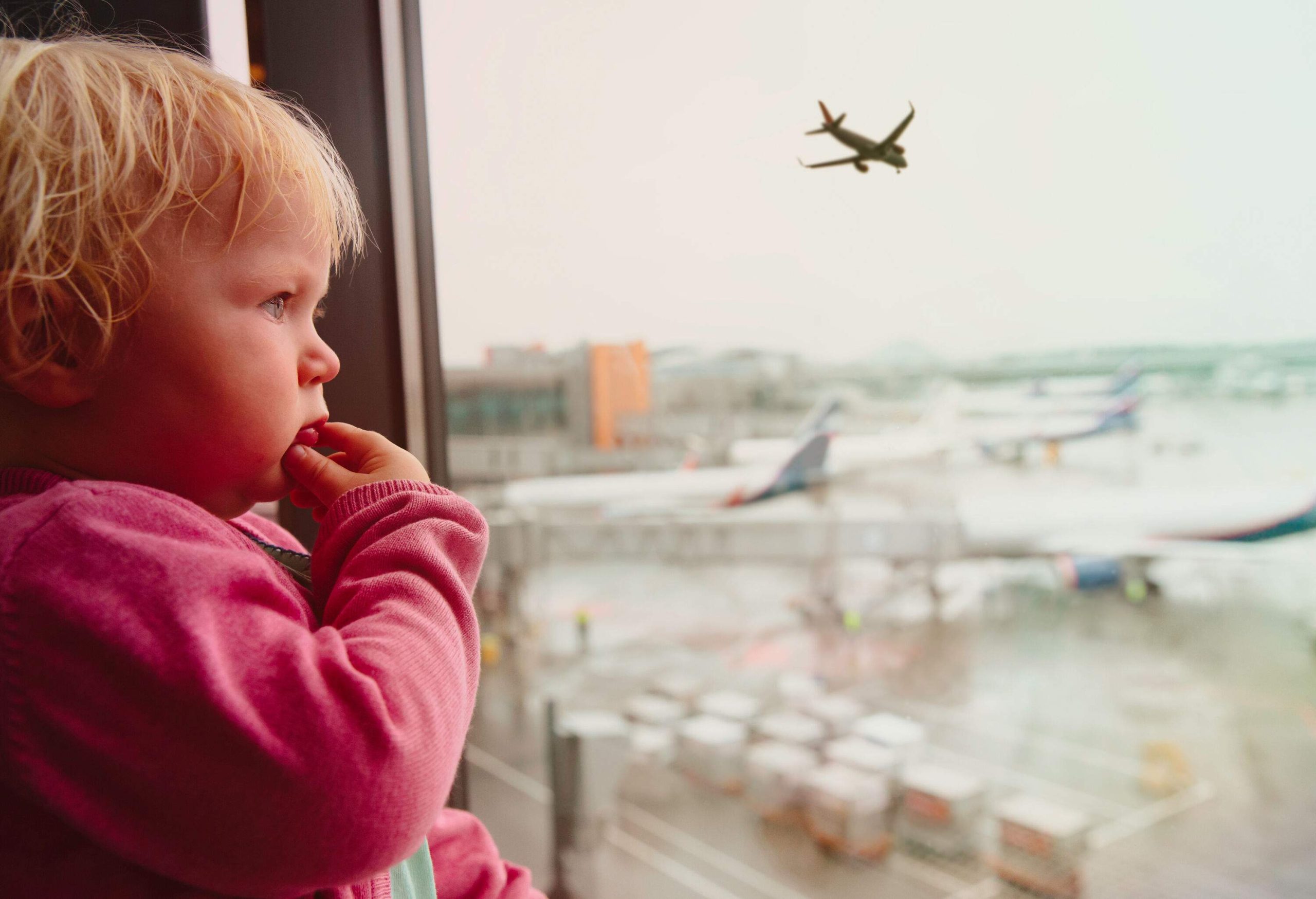 A baby wearing a pink sweater peers out of a window as a plane flies overhead.