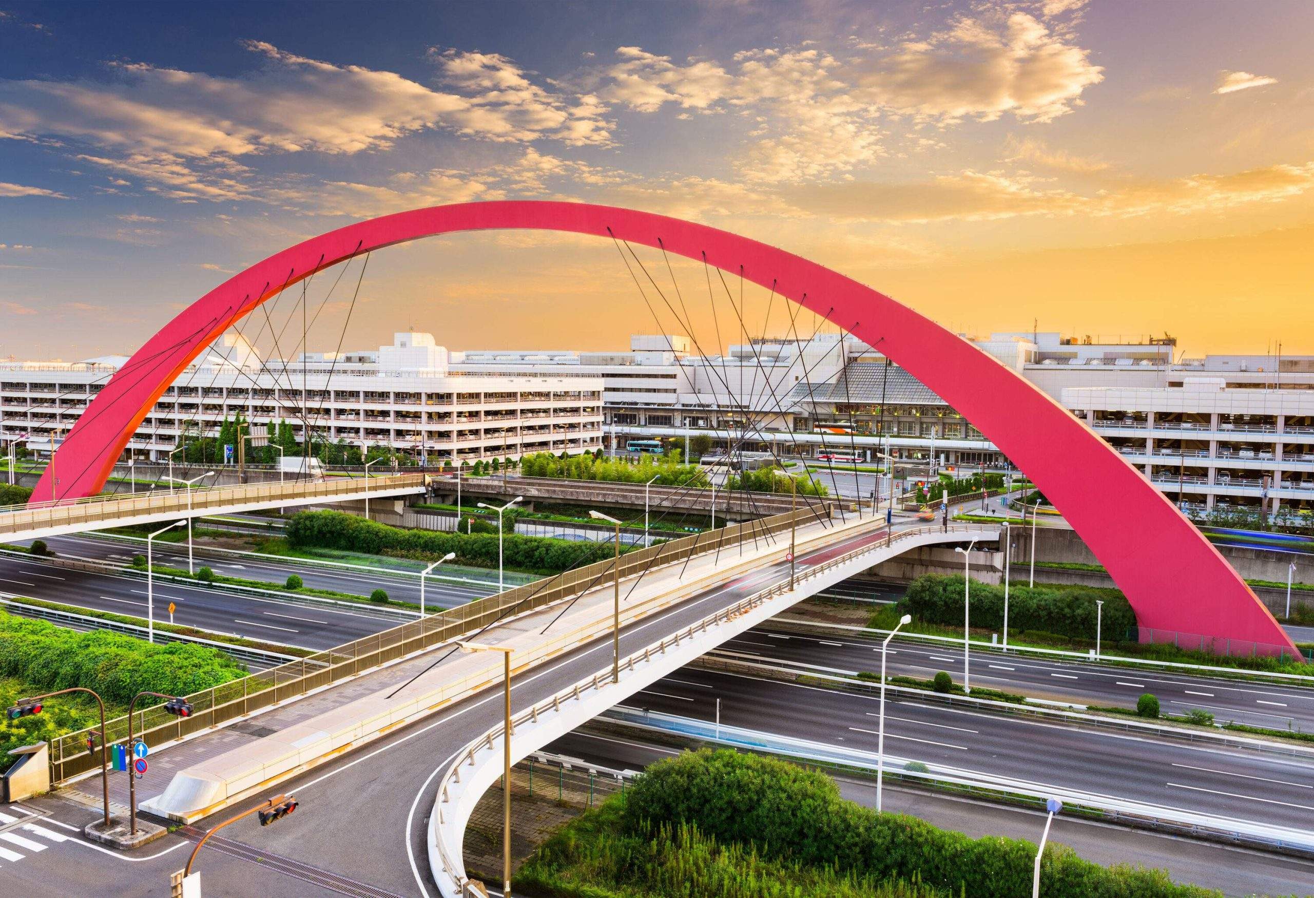 Two road bridges with an aesthetic design suspended by cables in a red sky arch alongside the airport buildings span above the highway.