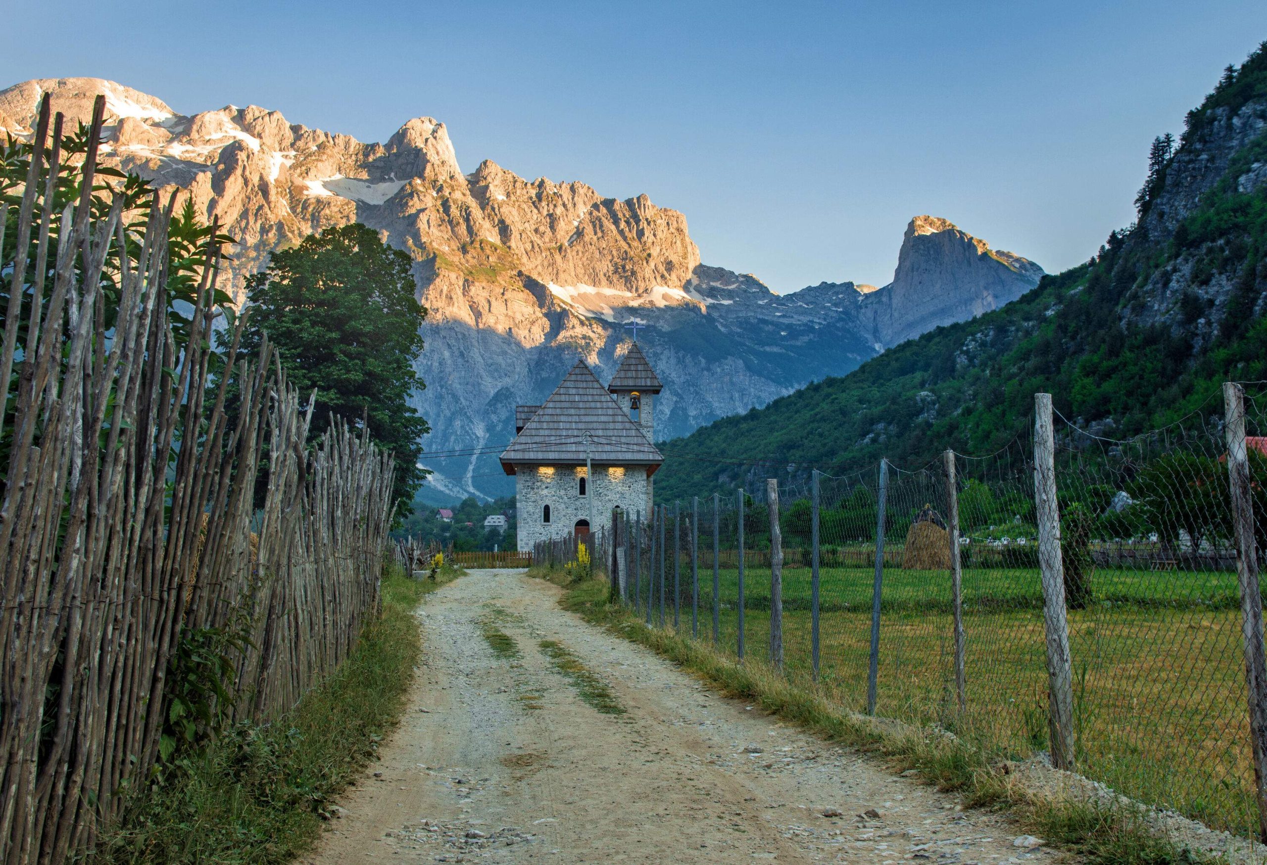 An unpaved road guarded by fences headed towards a tiny church with a steep rock mountain in the background.