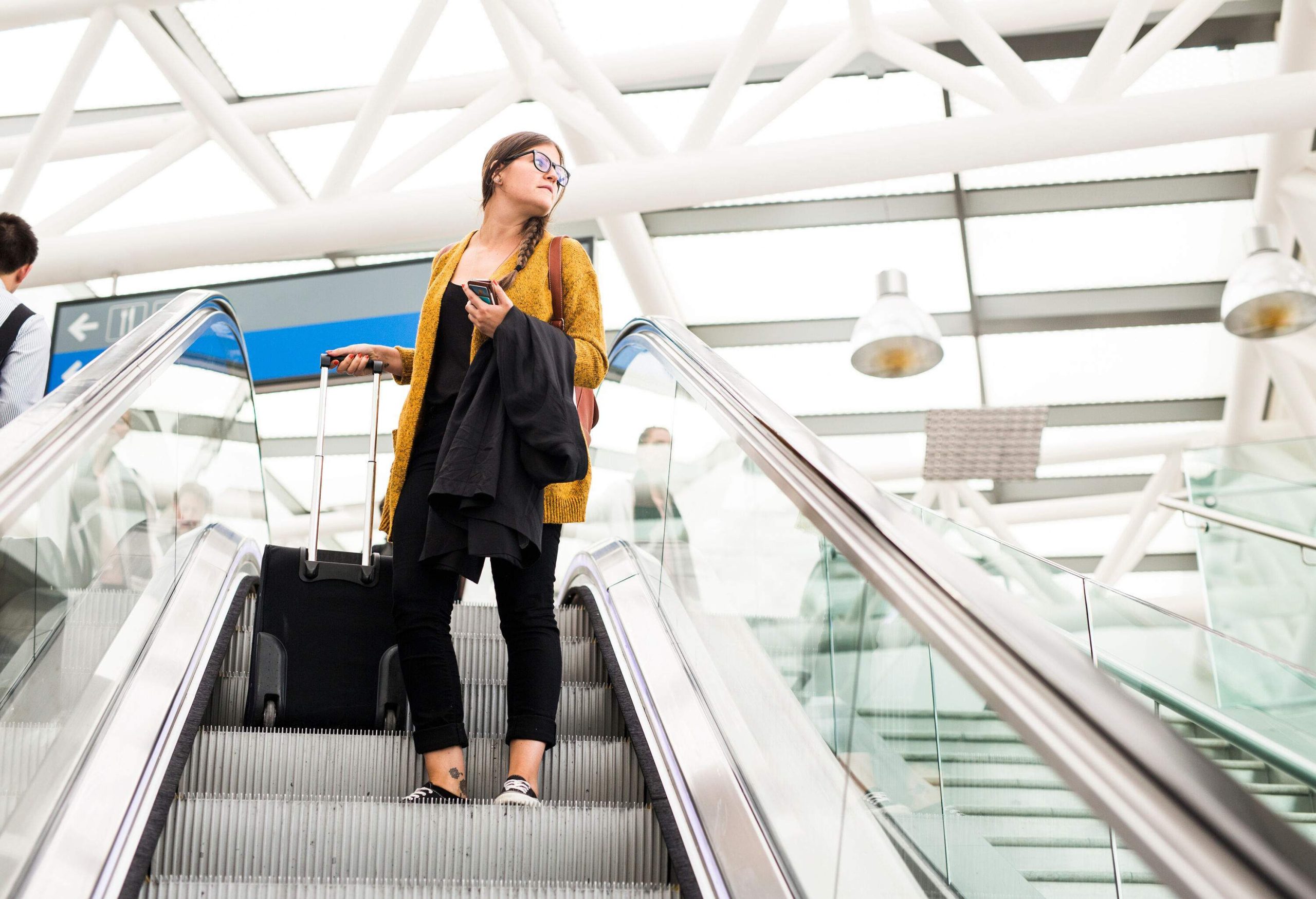 A woman with a suitcase standing on an escalator, distractedly gazing someplace.