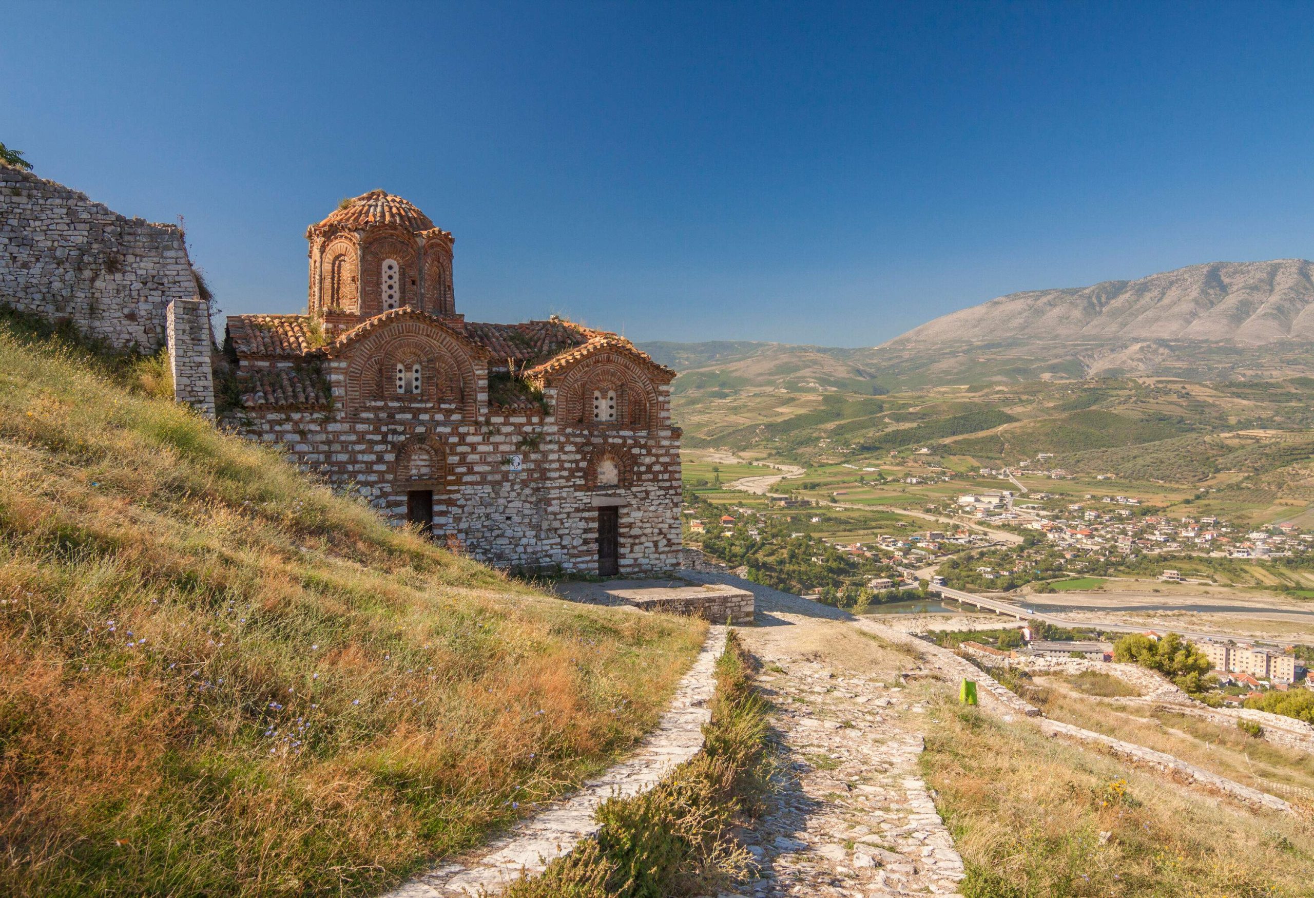 A stone church with dome roof perched on a grassy hillside and overlooking views of a town.