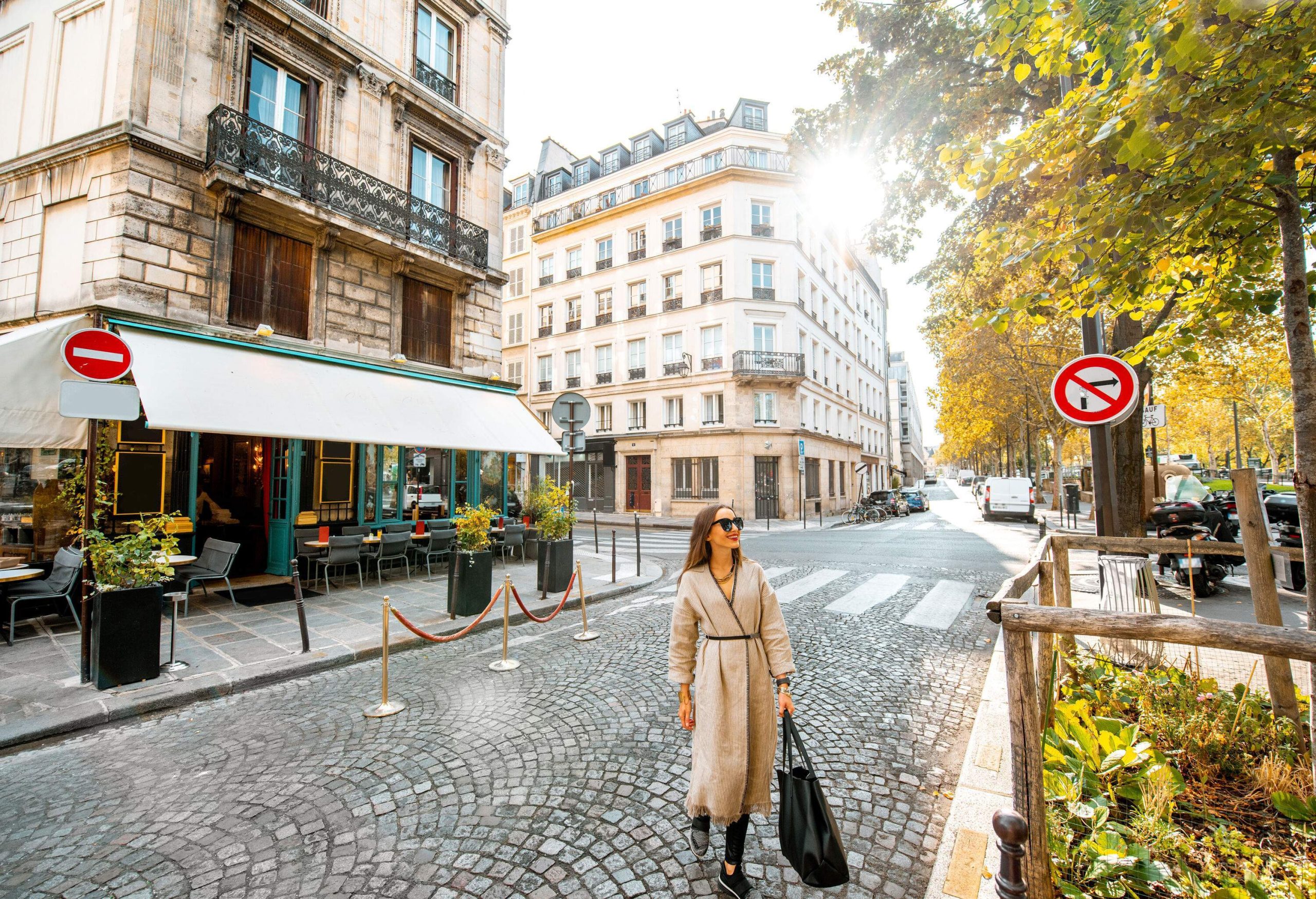 A stylish woman confidently crosses the street, with the charming facade of a restaurant building in the background, as a tree-lined street walk enhances the scene.