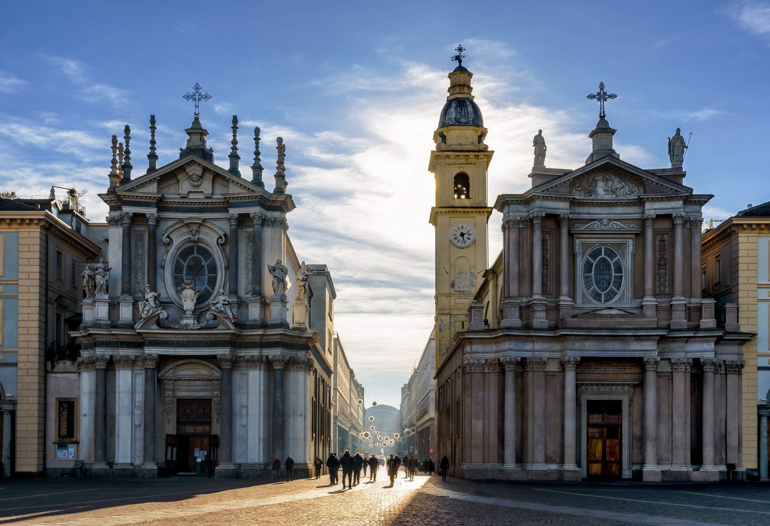 Many walk on the sunlit road in the middle of buildings lined along two adjacent churches on a cobbled square.