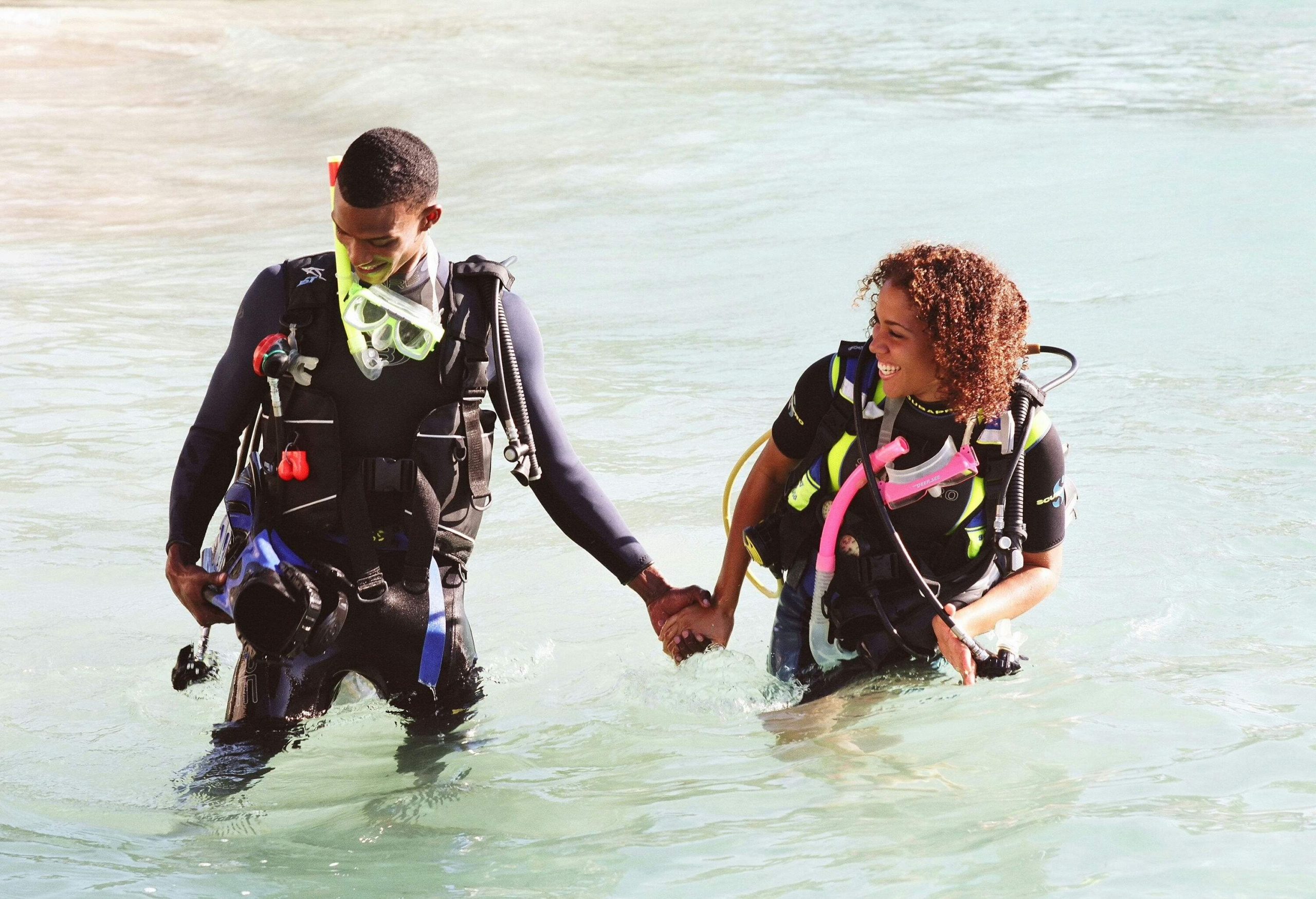 A couple wearing wetsuits and carrying snorkelling equipment wading in the water holding hands.
