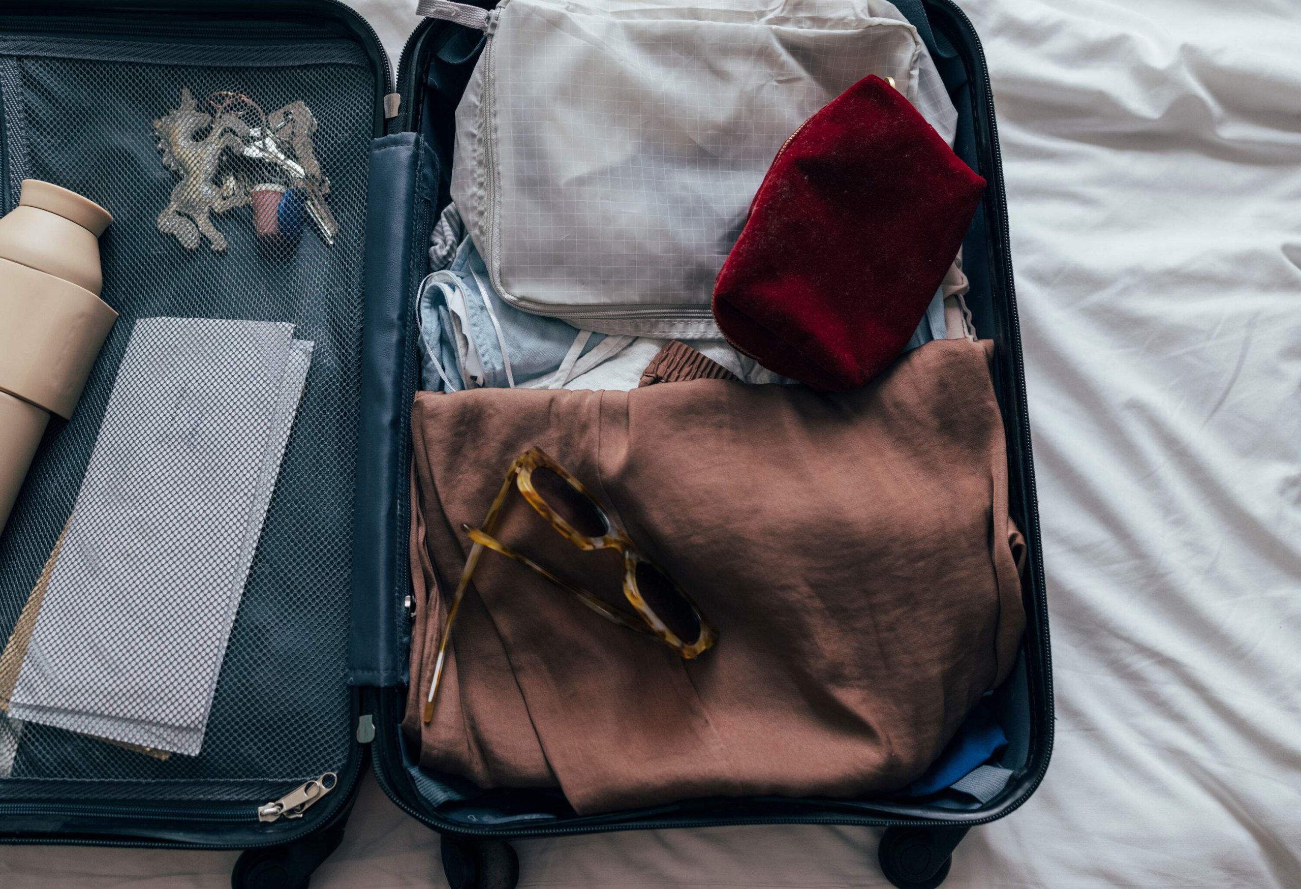 A cropped close-up of a grey spinner luggage from above on the bed on a holiday or a business trip containing sunglasses, a cosmetic bag, a water bottle, keys, clothes and plane tickets