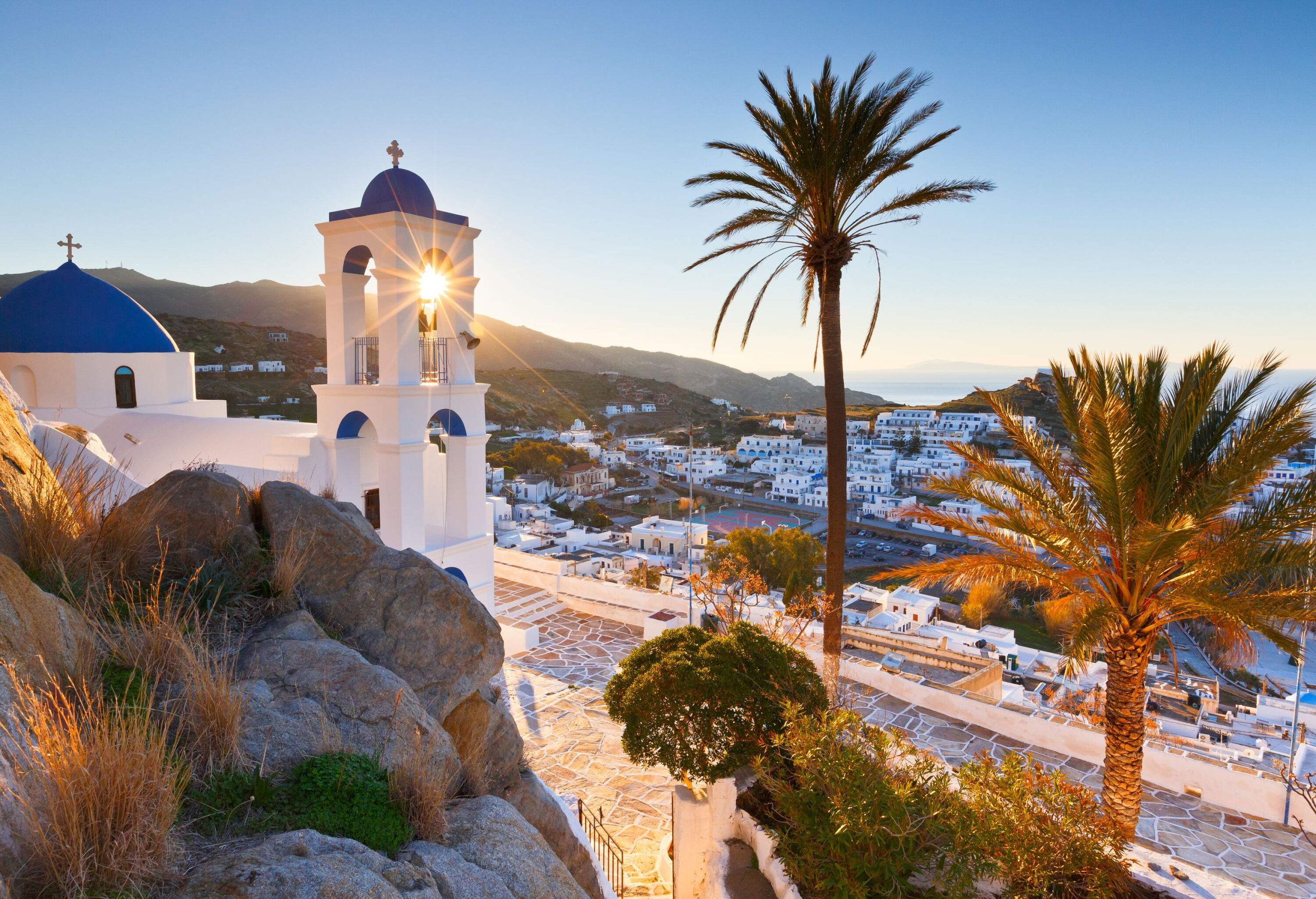 The sun shines through the belfry of a white hillside church with a blue dome roof and the surrounding trees and buildings.