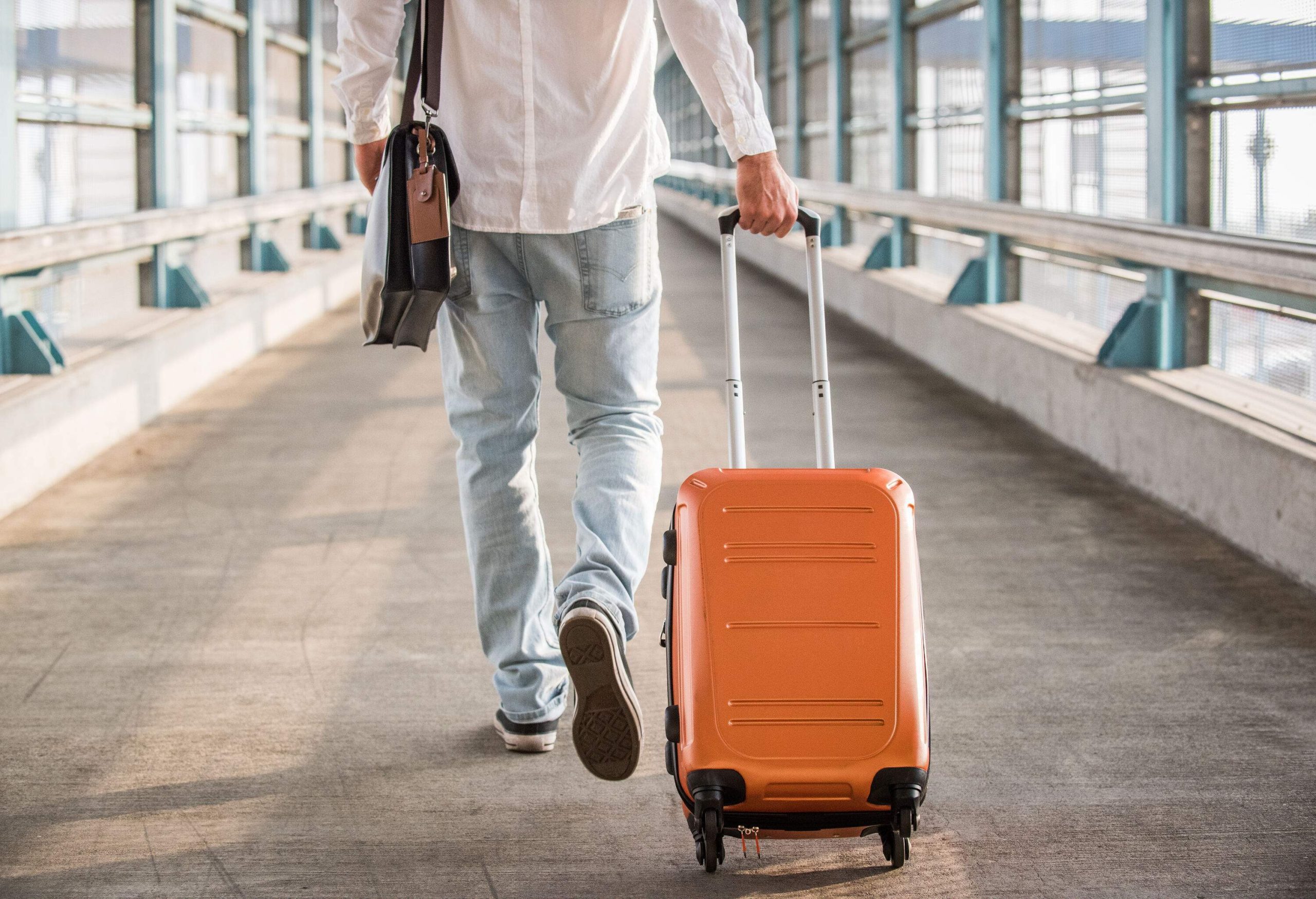 A man wearing casual attire, carrying a backpack and dragging a suitcase, walks along a walkway leading to a railway station.