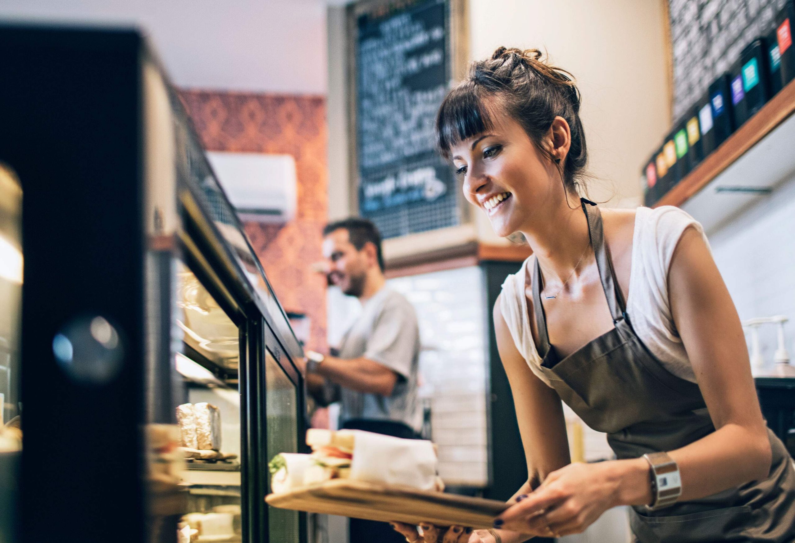 A brunette woman in an apron places sandwiches on a restaurant counter display.