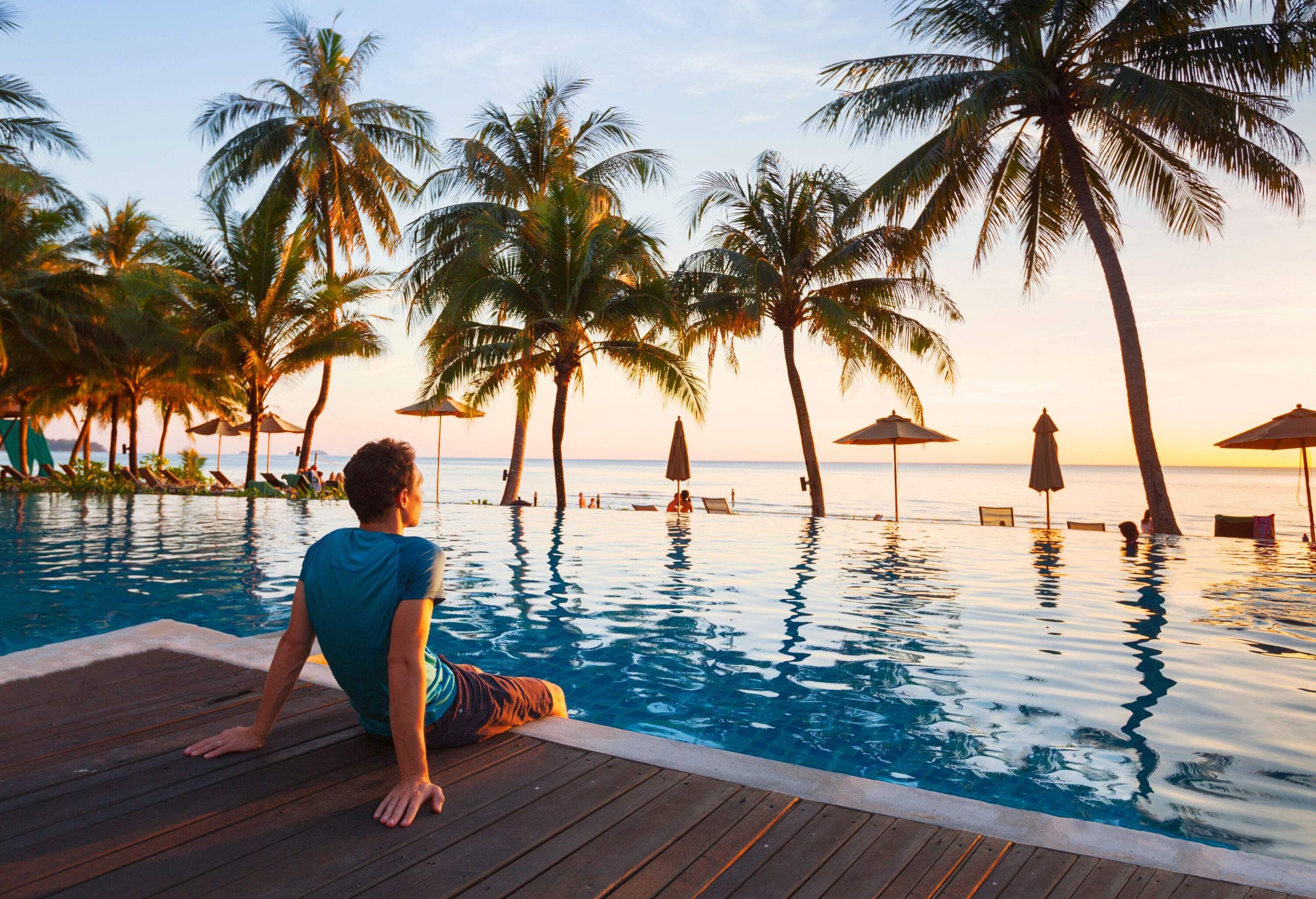 A lone man sits on the wooden deck of an infinity pool overlooking the sea at sunset.