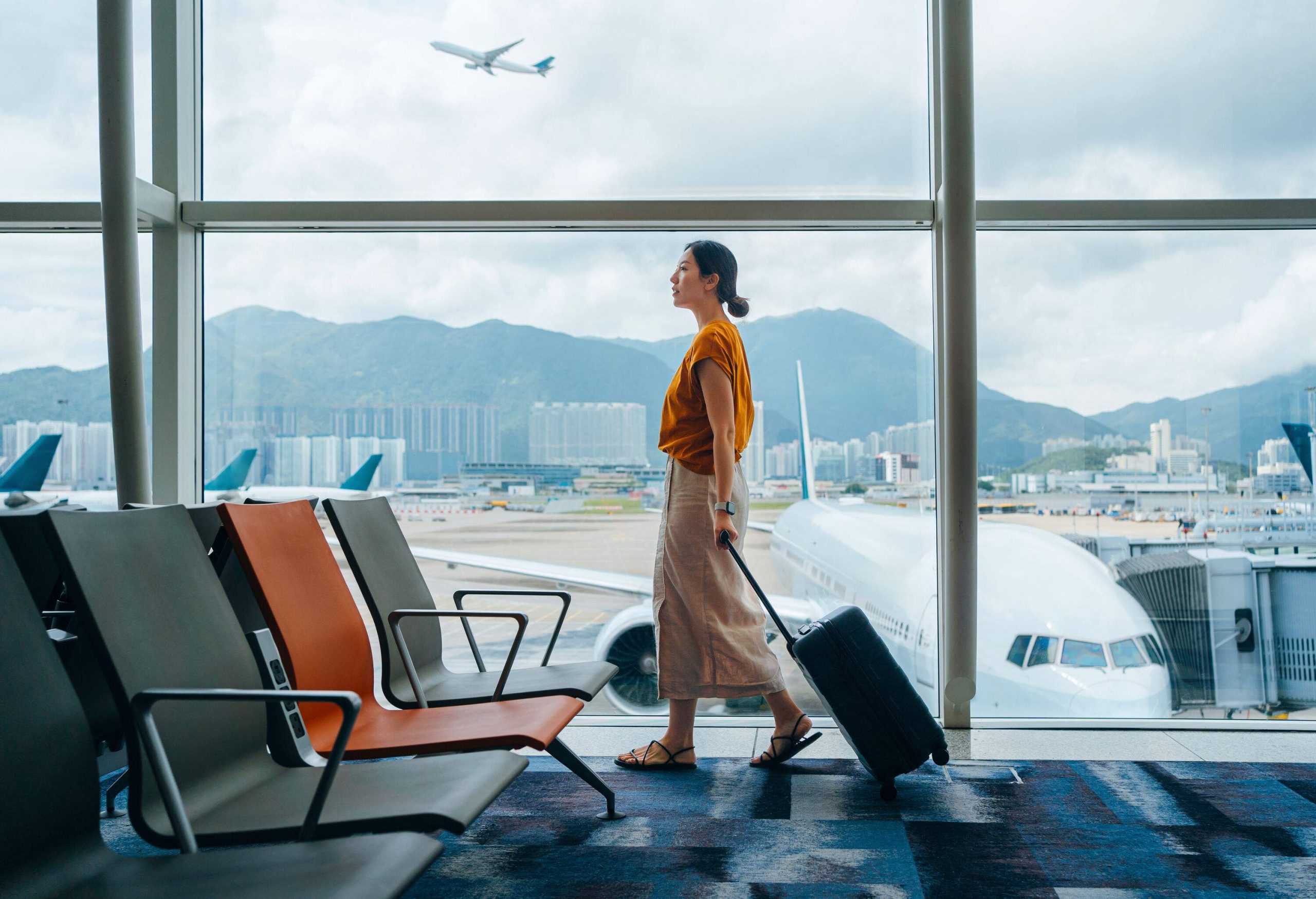 Young Asian woman carrying suitcase, walking by the window at airport terminal. Young Asian female traveller waiting for boarding at airport. Business travel. Travel and vacation concept