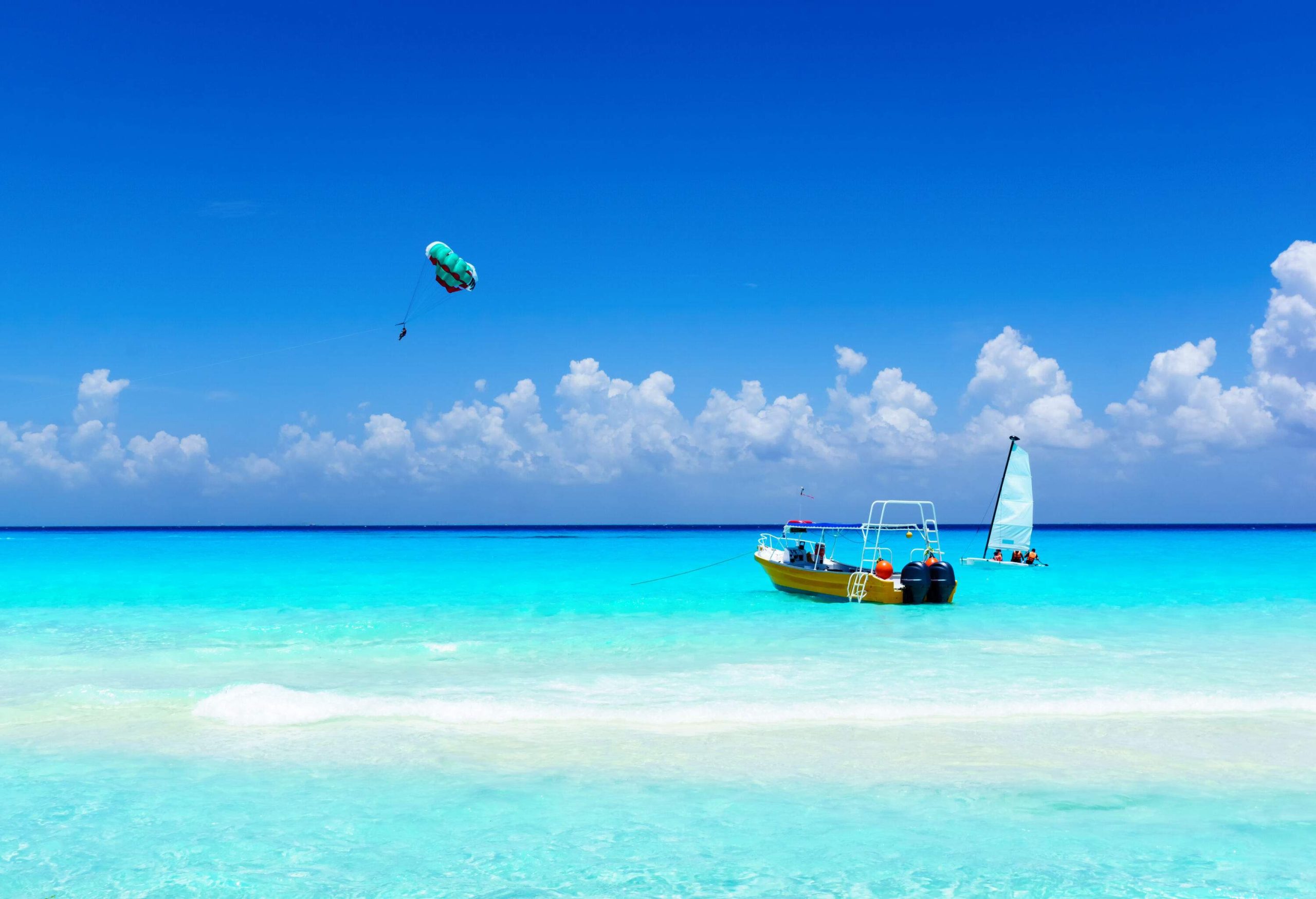 A sailboat and an anchored boat on a pristine beach with distant views of a parasailer.