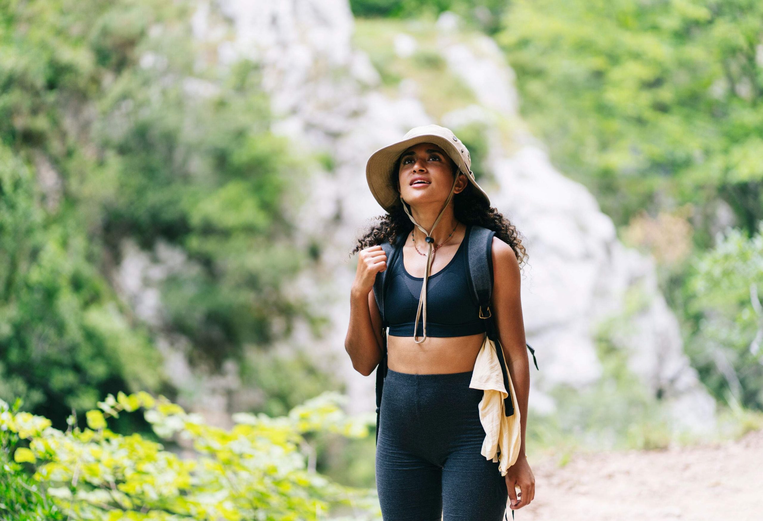 A woman in a sports bra with black leggings and a brown hat is looking up into the forest.