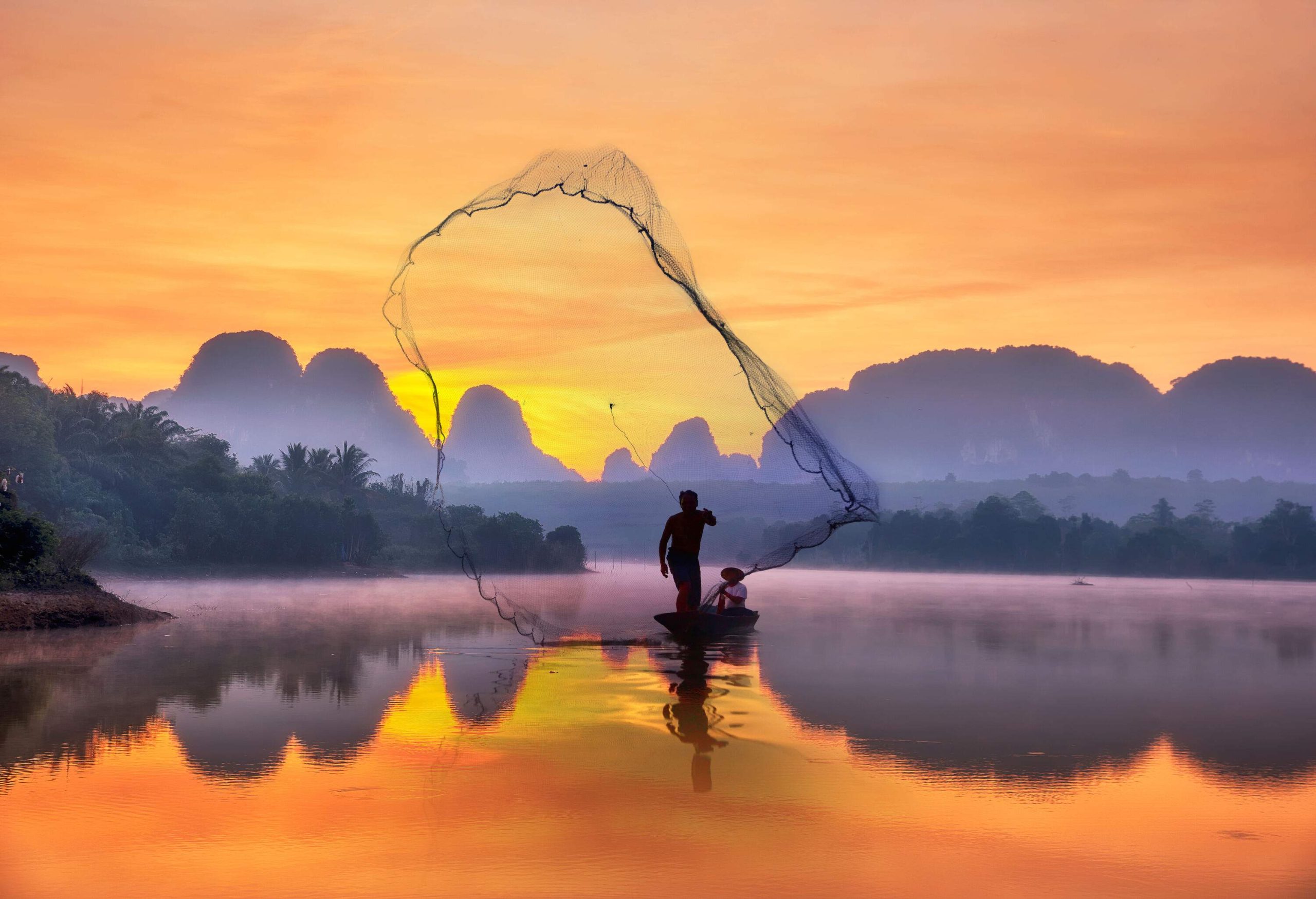 A fisherman on a boat throws a fishing net to a calm river that reflects the surrounding misty mountains and orange skies.