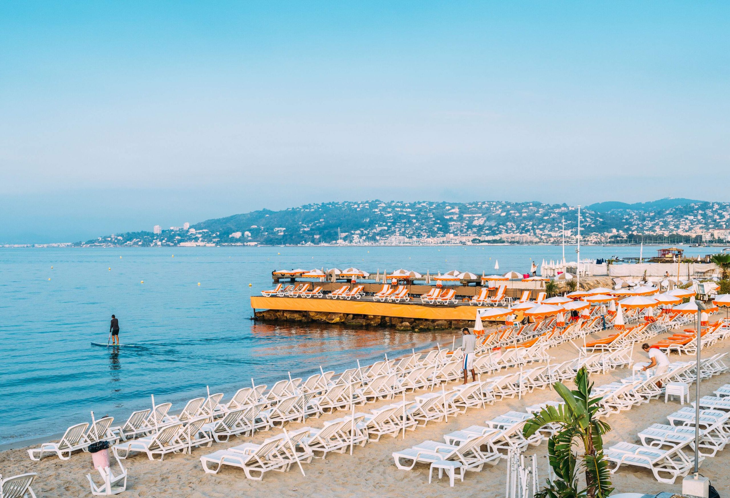 A beach with rows of sunbeds and a person paddleboarding on the ocean.