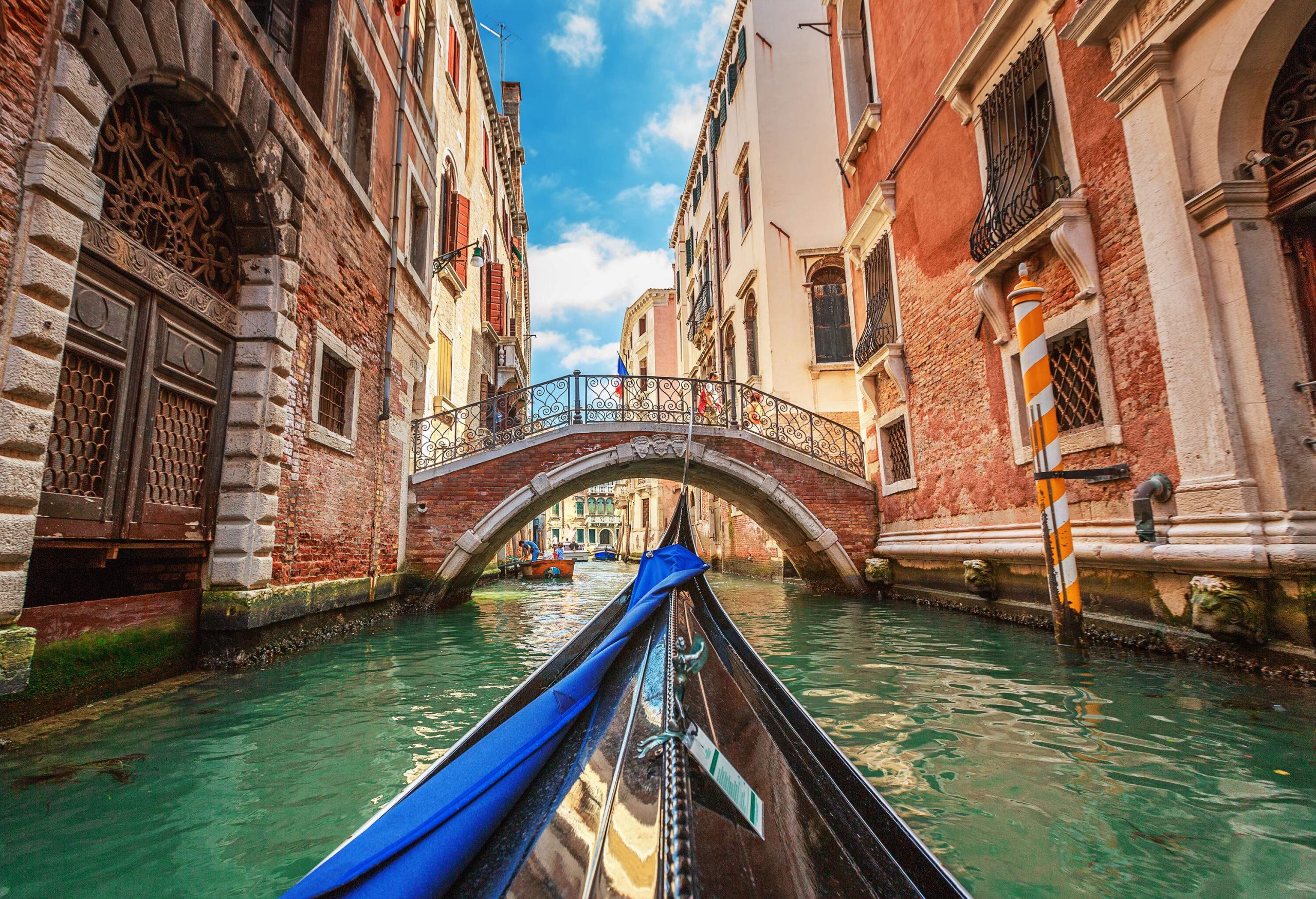 A gondola cruises a water canal as it approach a bridge between tall buildings.