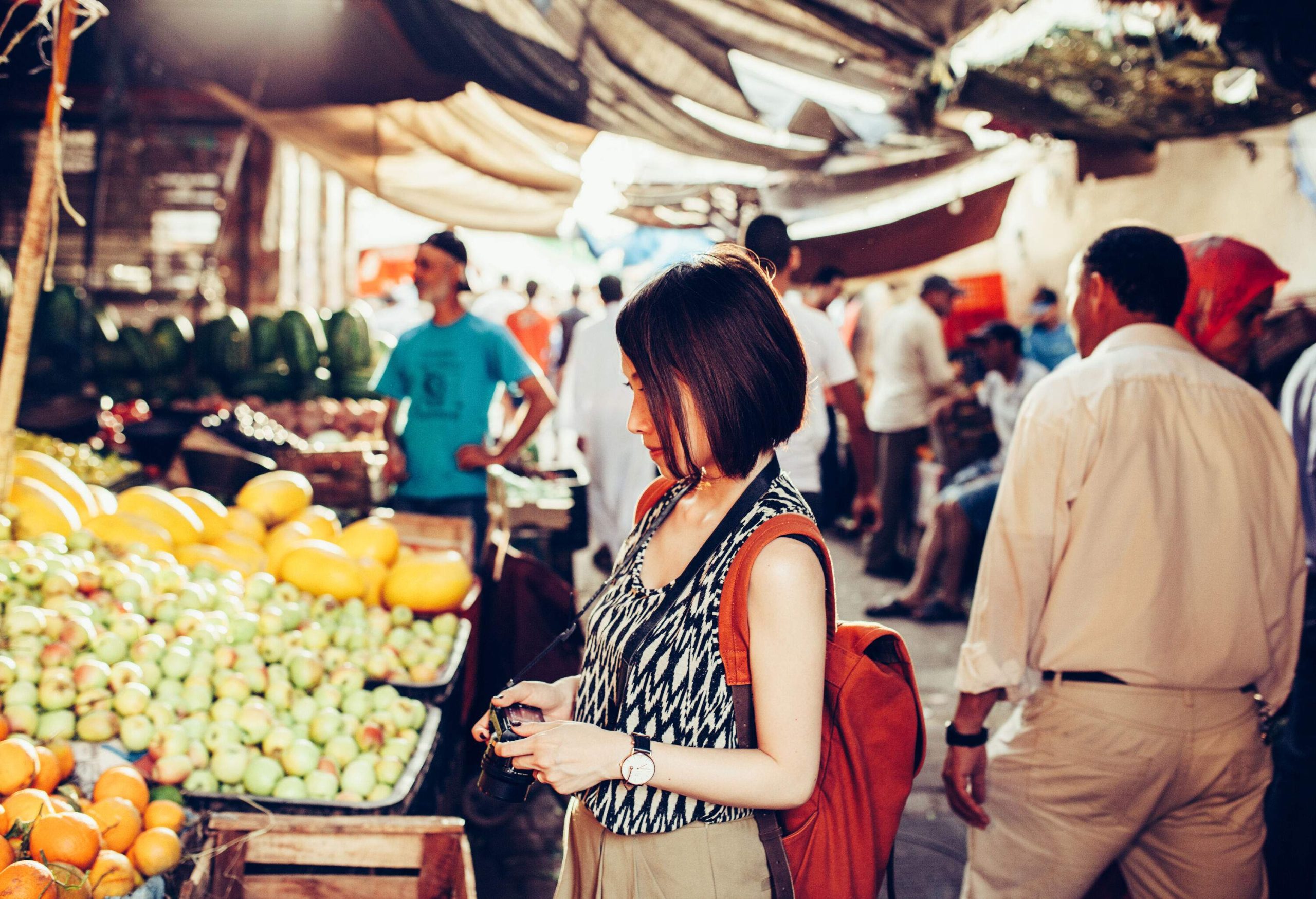 A young tourist holding a digital camera standing in front of a fruit stall.