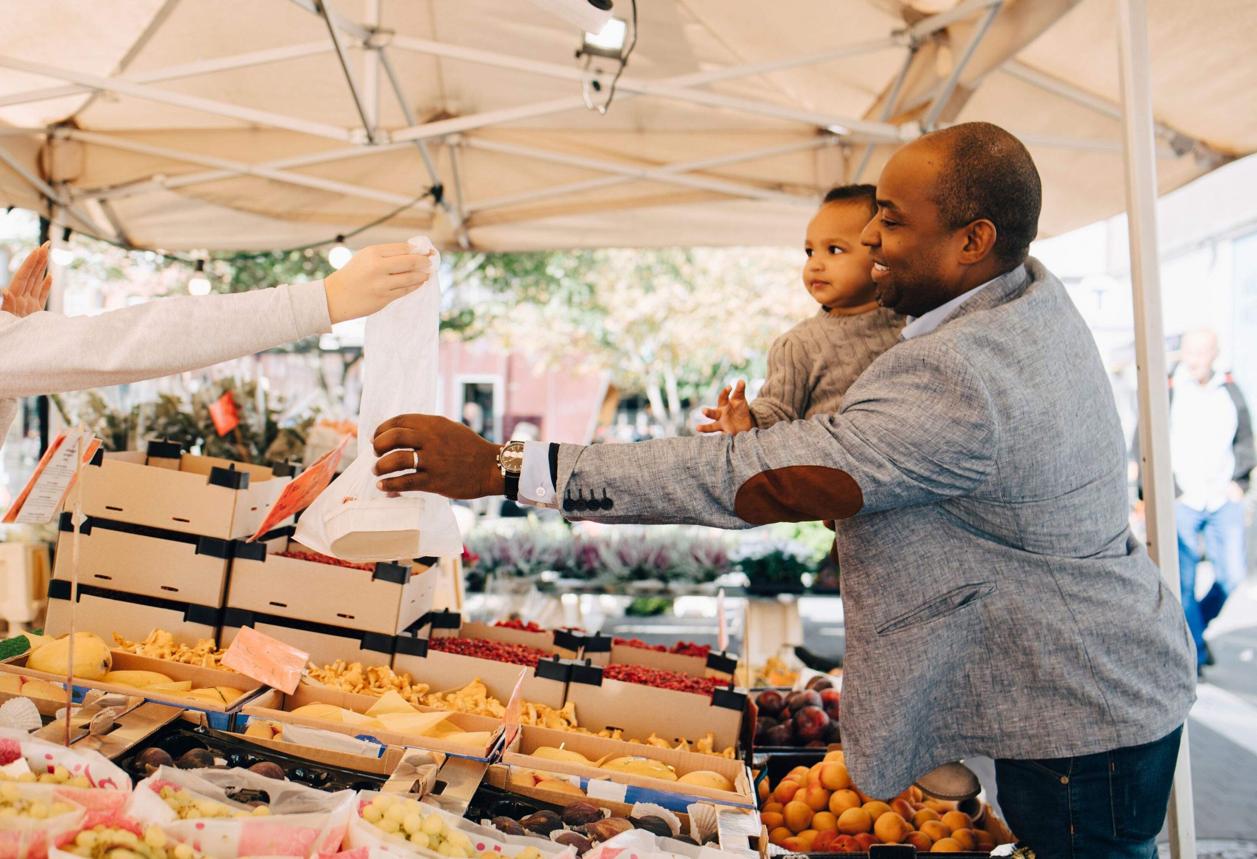 A person carrying a little child while reaching for a plastic bag of merchandise at a fruit shop.