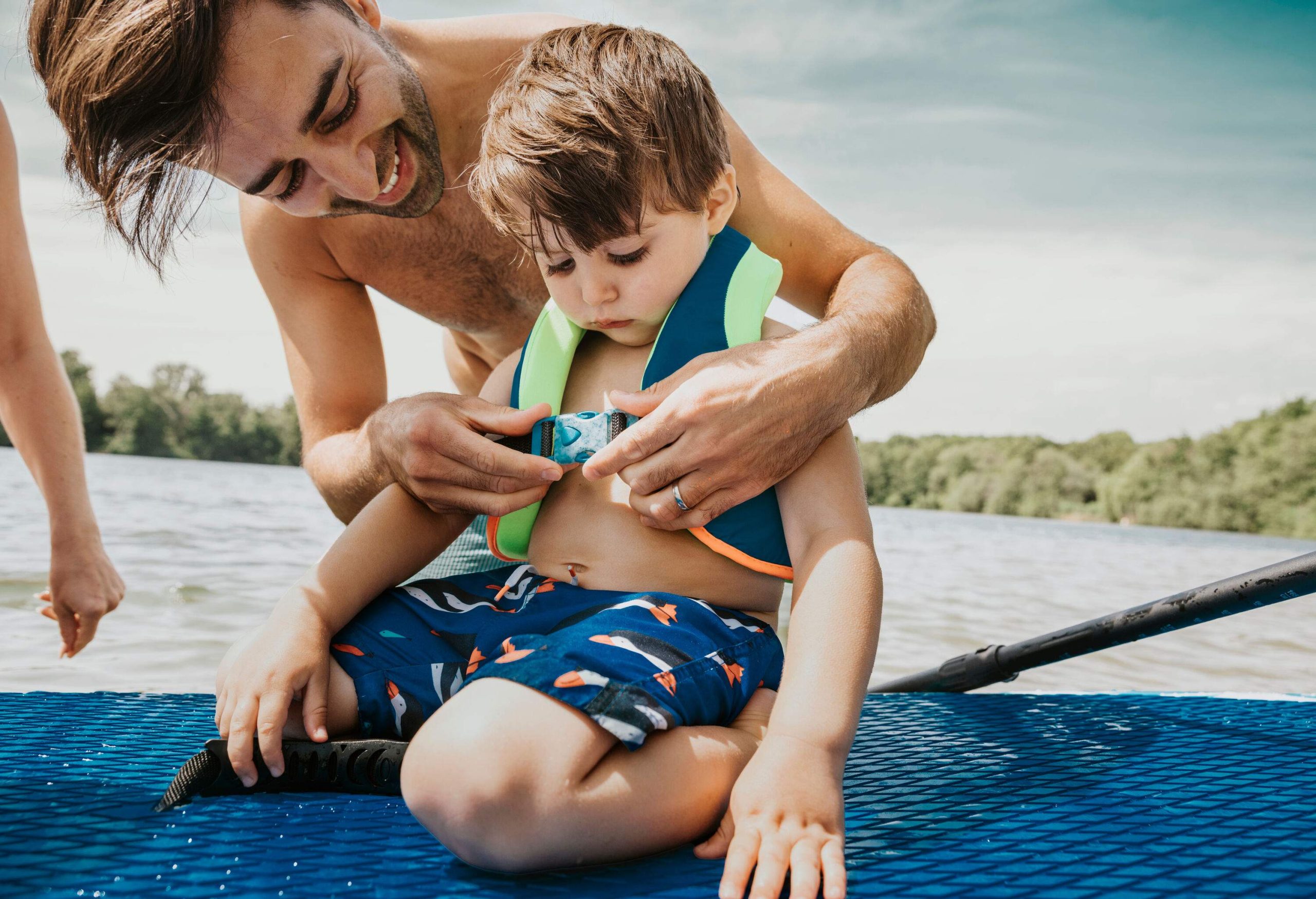 A young man putting a float vest on a little boy sitting on a paddleboard.