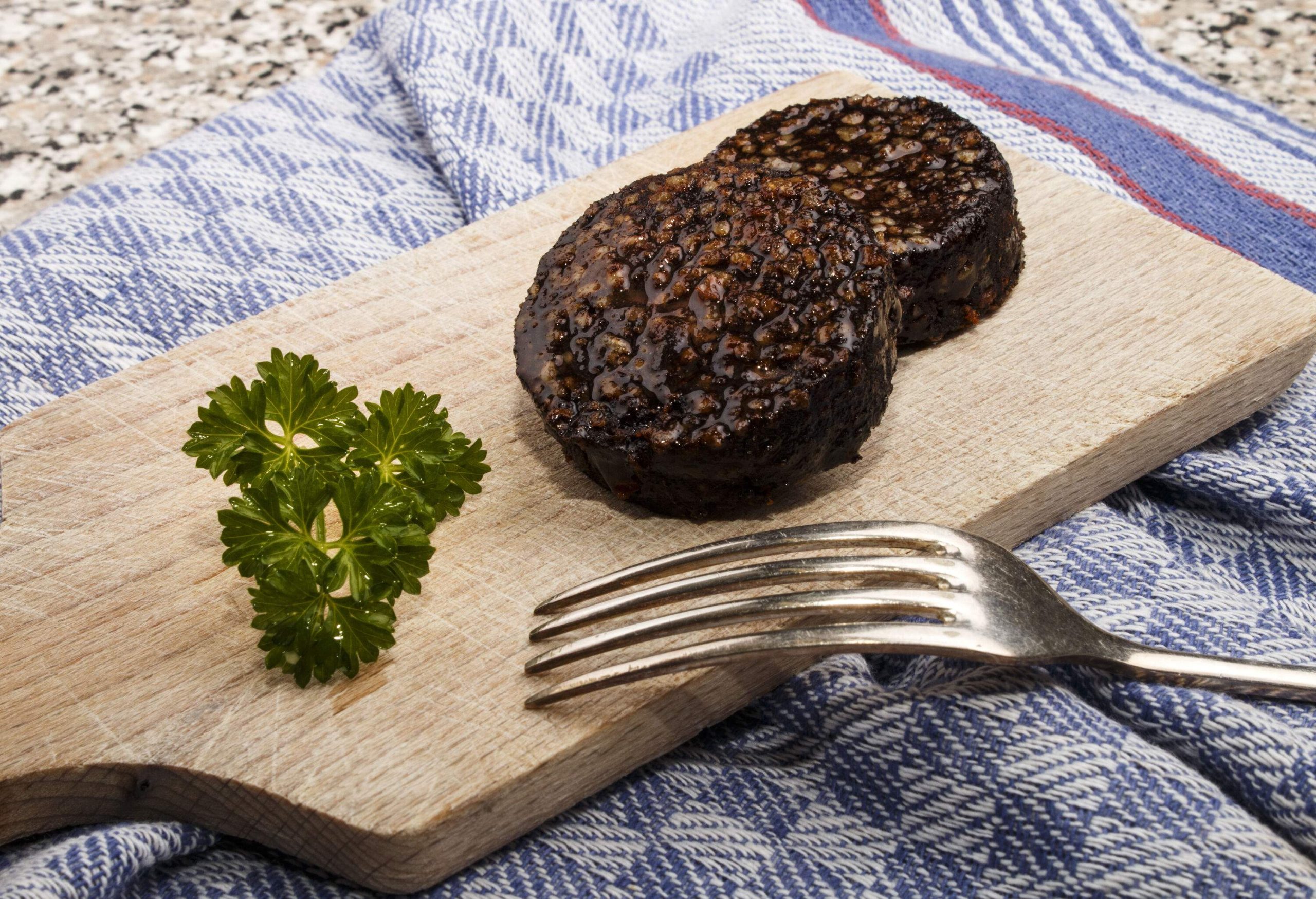 Two slices of black pudding on a wooden board with herbs and a fork.