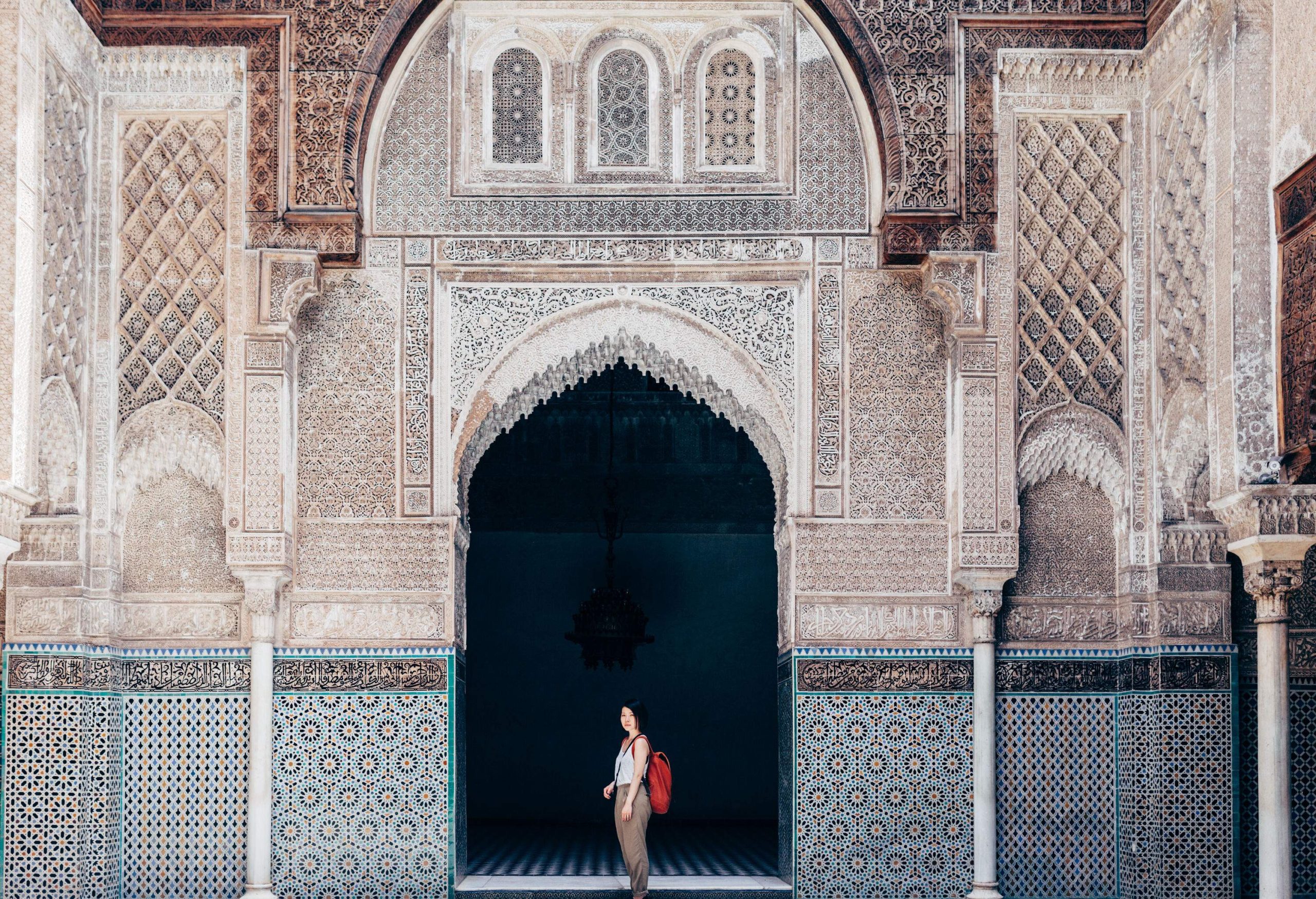 A woman stands under a building's arch entrance with walls intricately carved with unique geometric patterns.