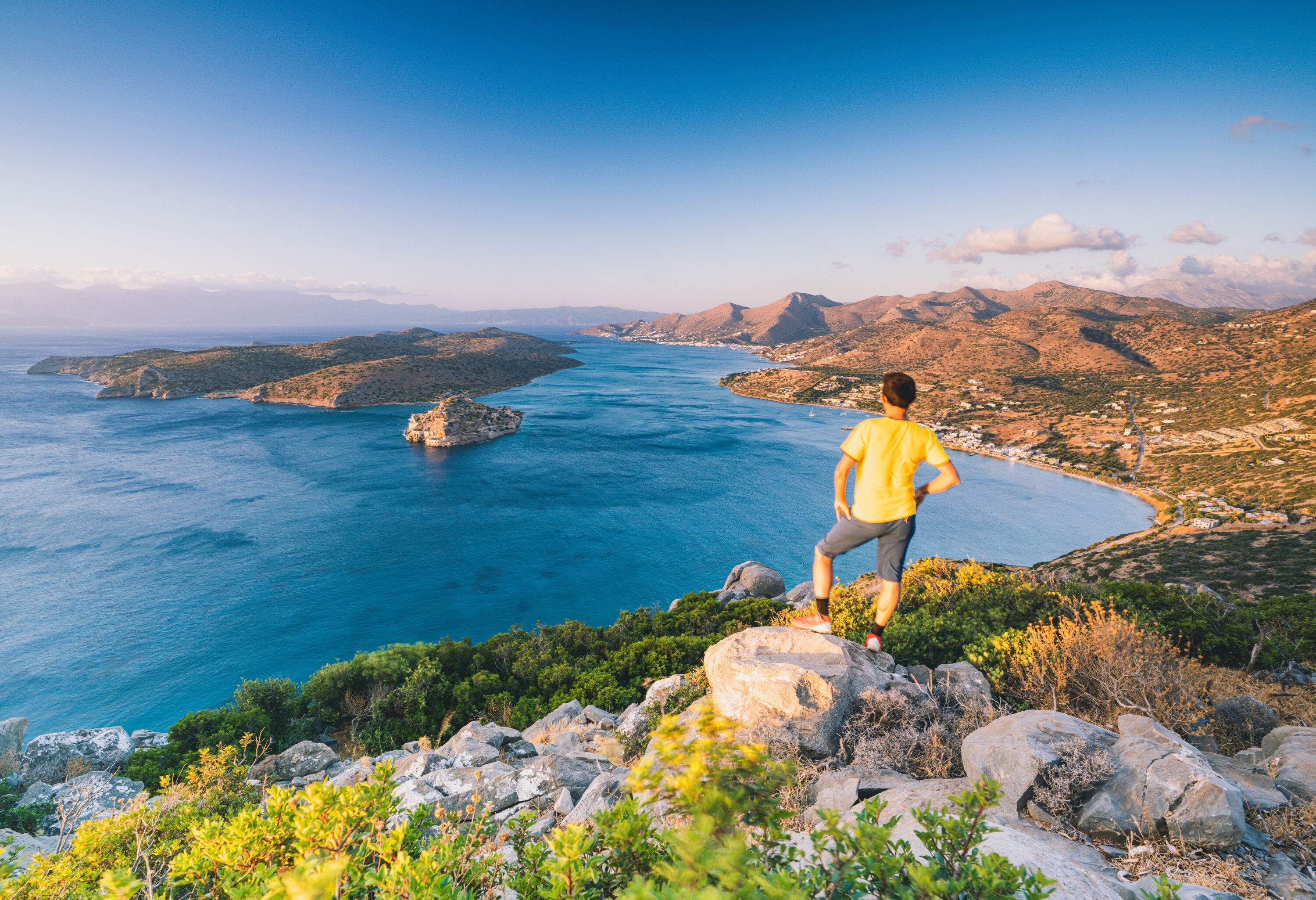 A man in a yellow shirt standing on a rock, taking in the spectacular view of a bay around an island.