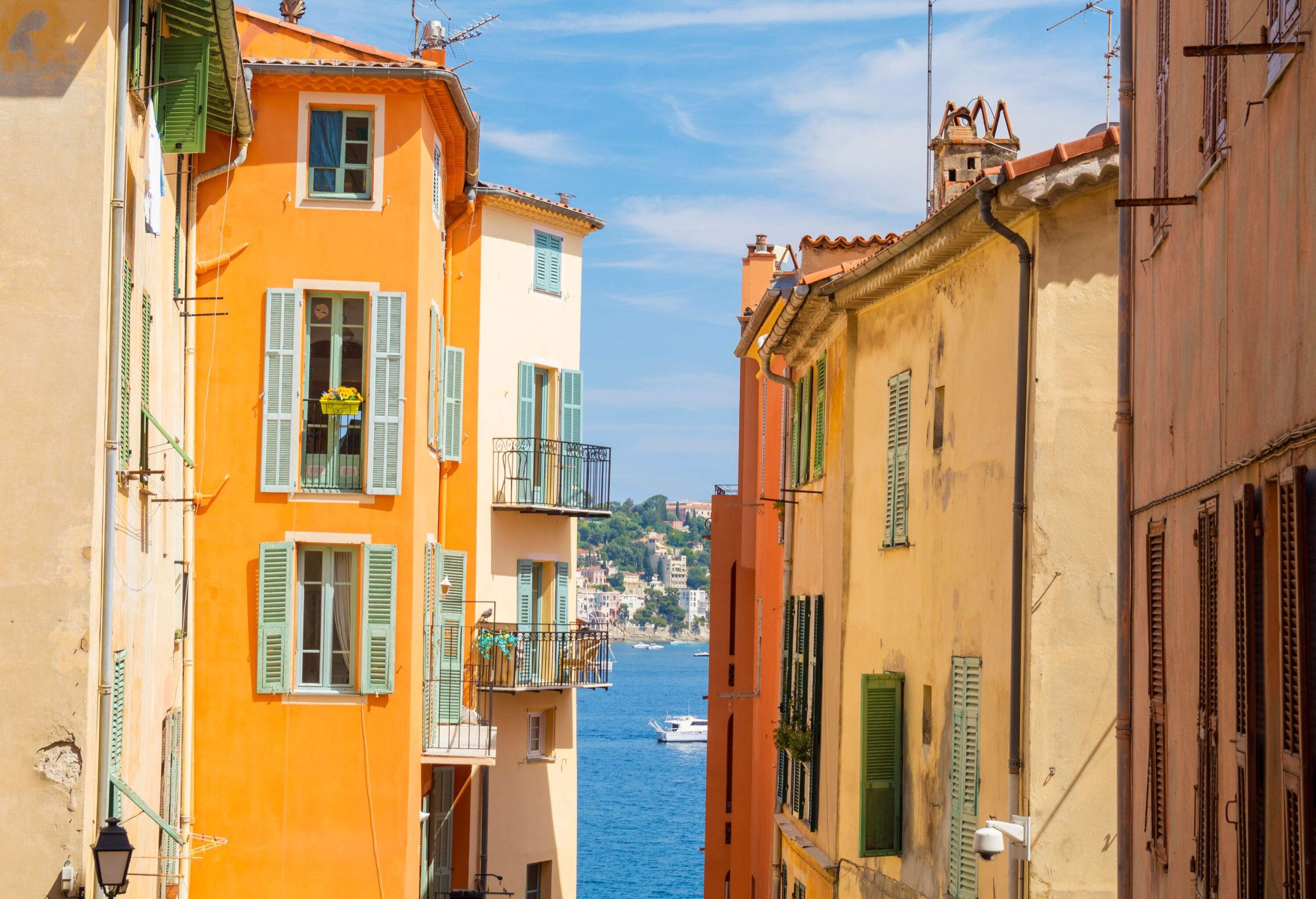 Between colourful buildings is a view of boats sailing on calm blue waters surrounded by a hill packed with structures and trees.