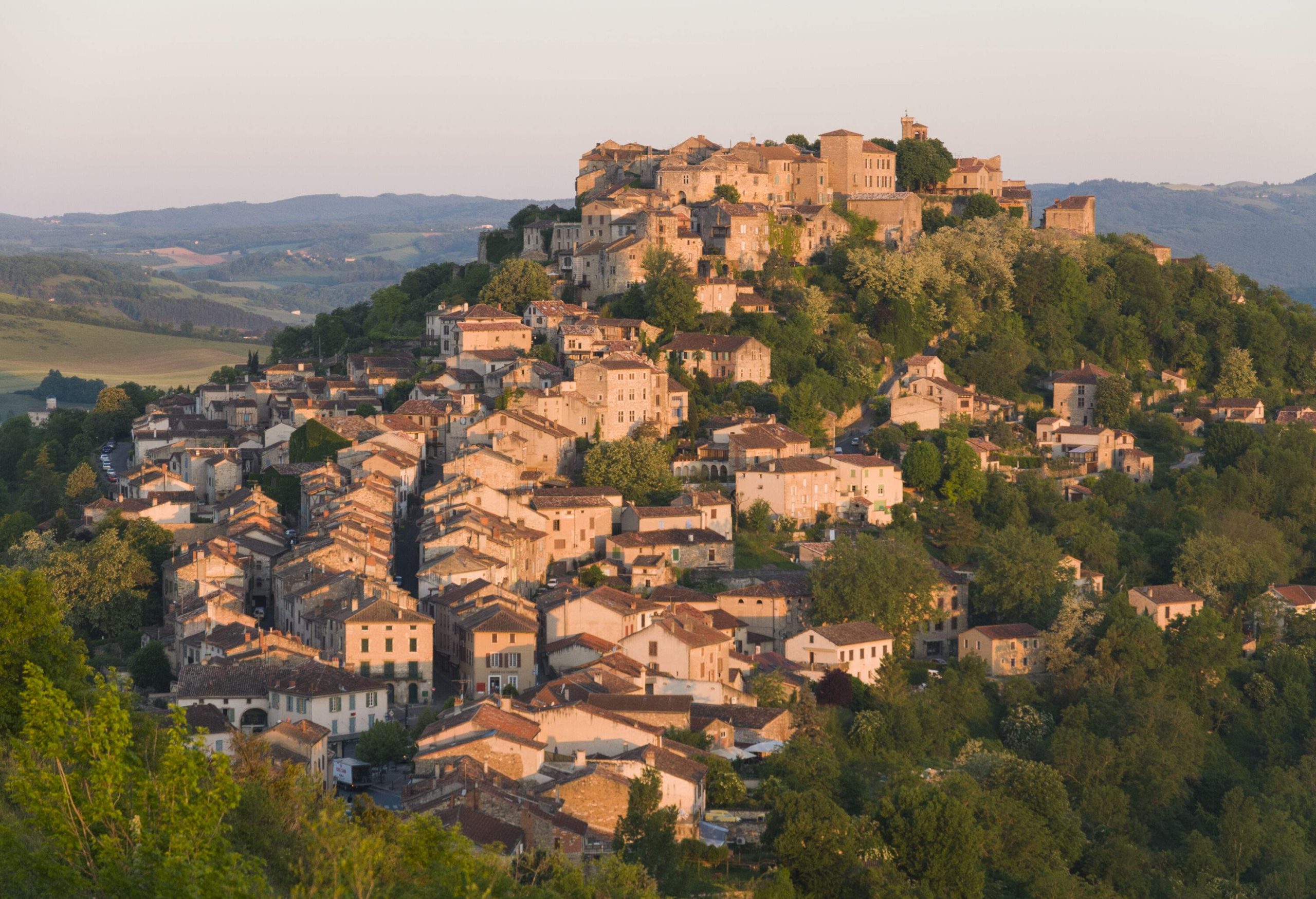 An old medieval town with classic stone buildings perched on a lush hilltop at sunrise.