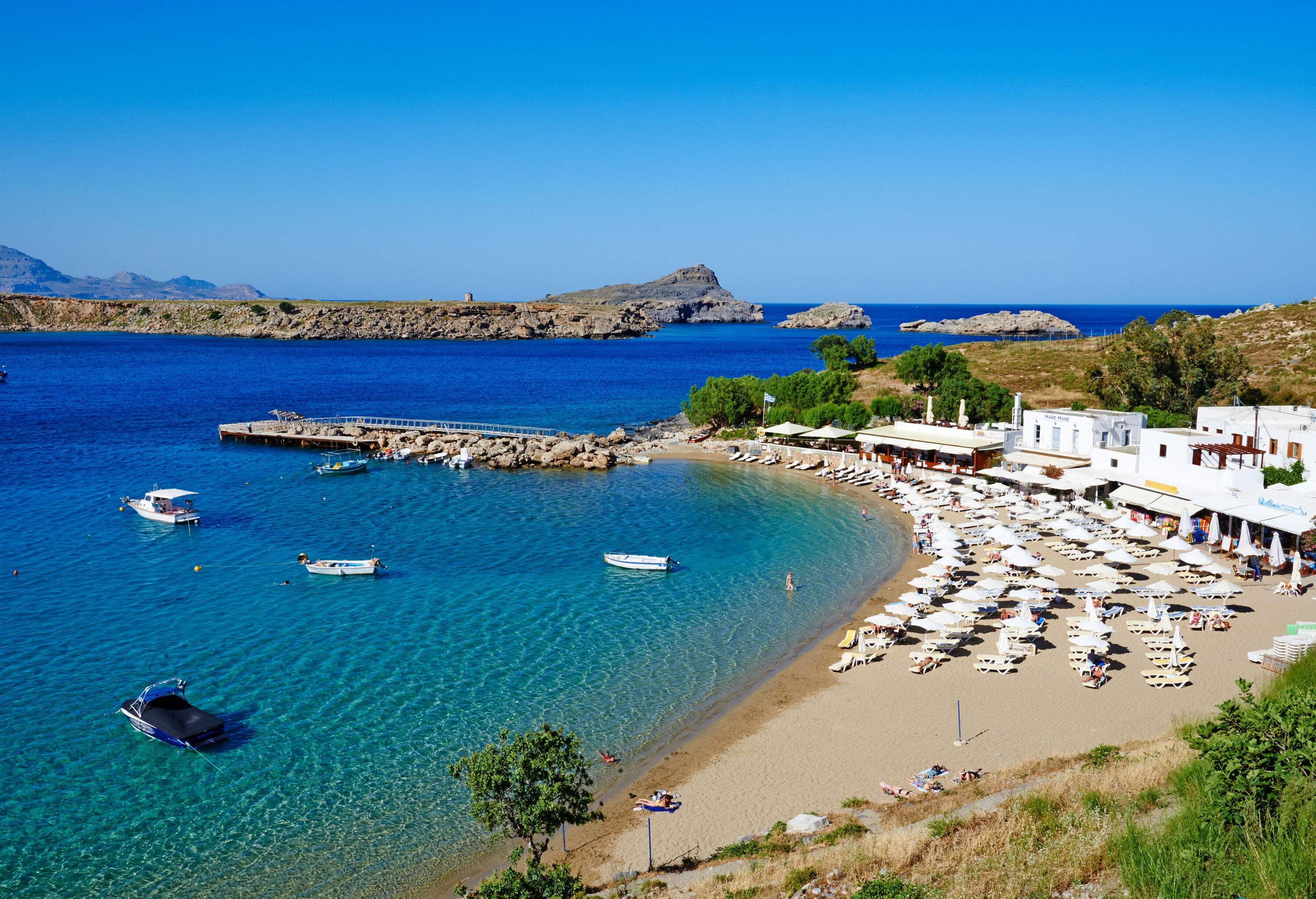 A beach with rows of sunbeds and umbrellas and anchored boats on a harbour. 