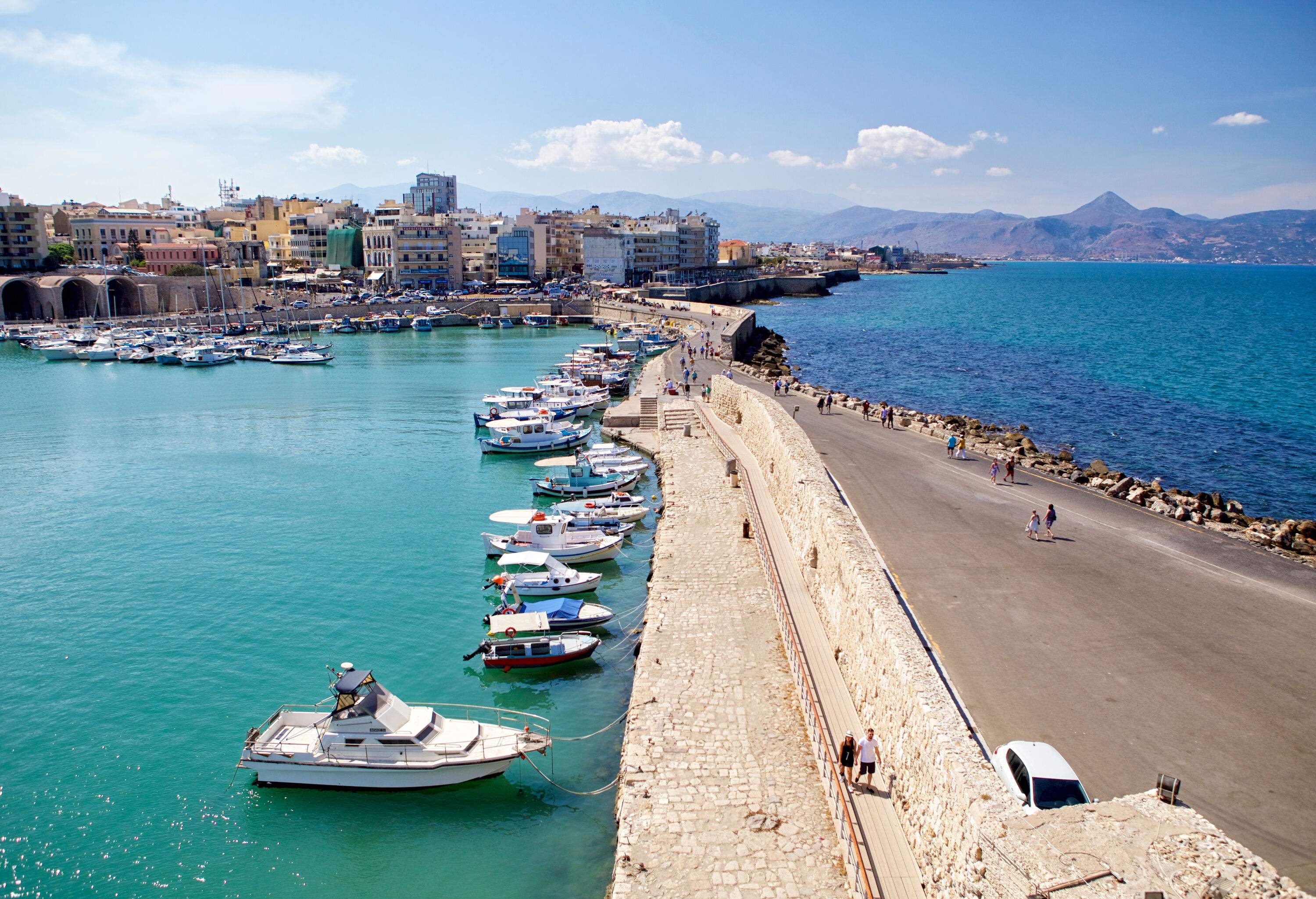 A lovely beachfront town overlooks a thriving harbour with many moored boats along a walled road, surrounded by dazzling waves.
