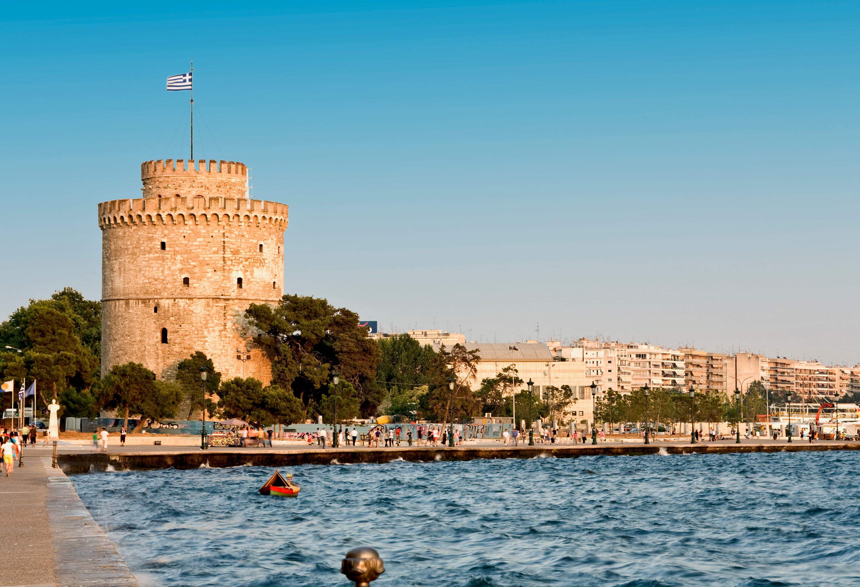 A white cylindrical tower with a Greece flag on a pole along a busy promenade.