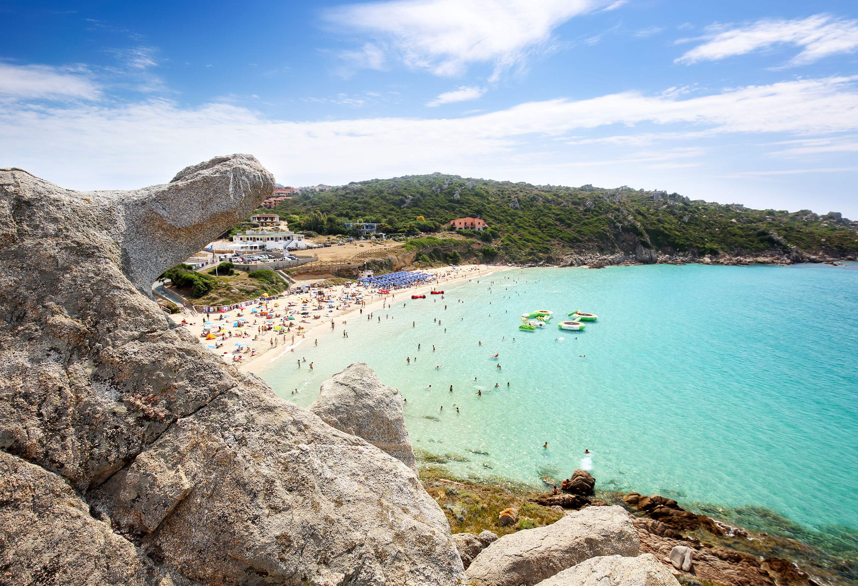 A bustling turquoise beach and a lush island are seen from the top of a rocky hill.
