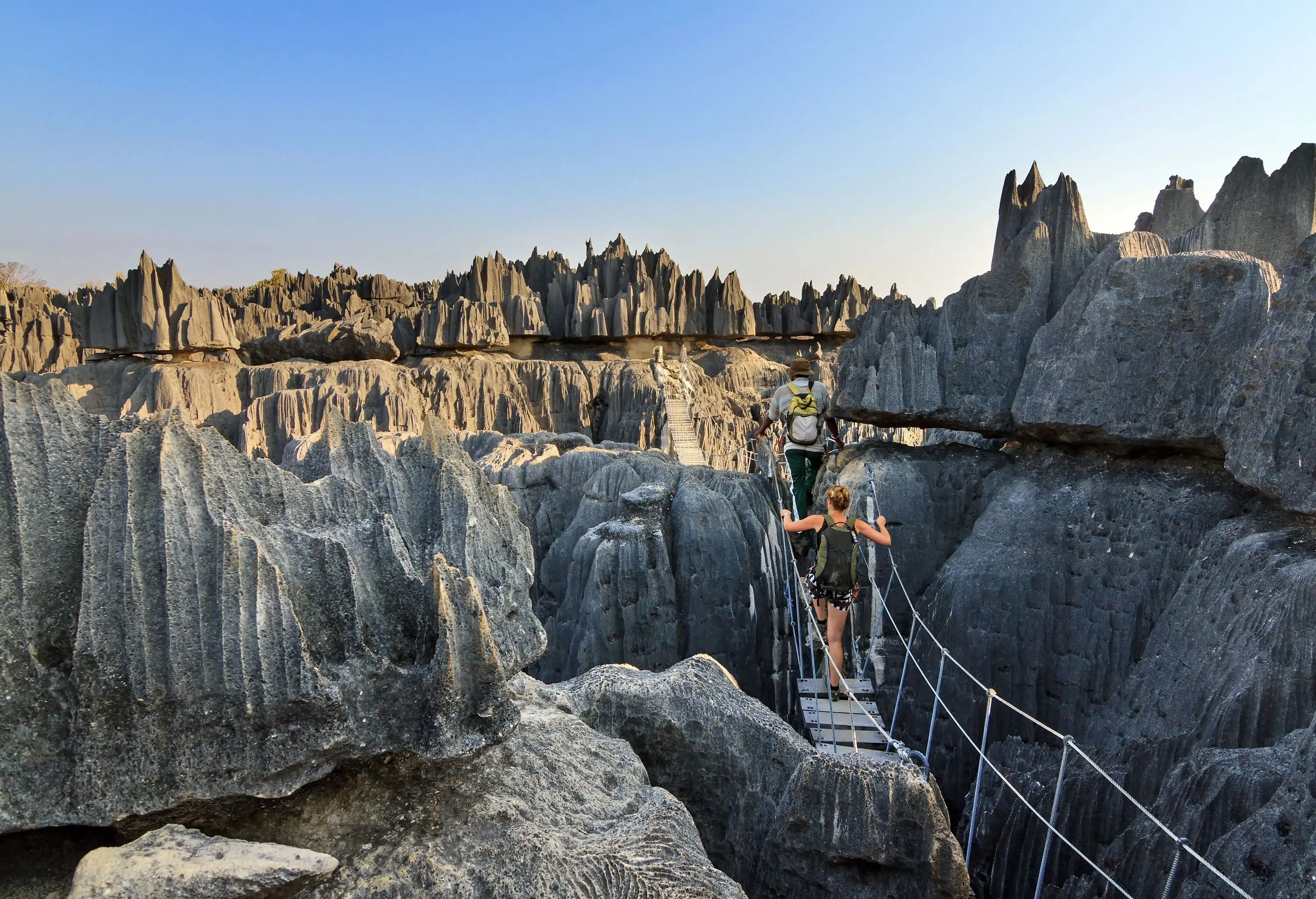 Two hikers cross a hanging bridge on the narrow cliffs of mountains with sharp peaks.